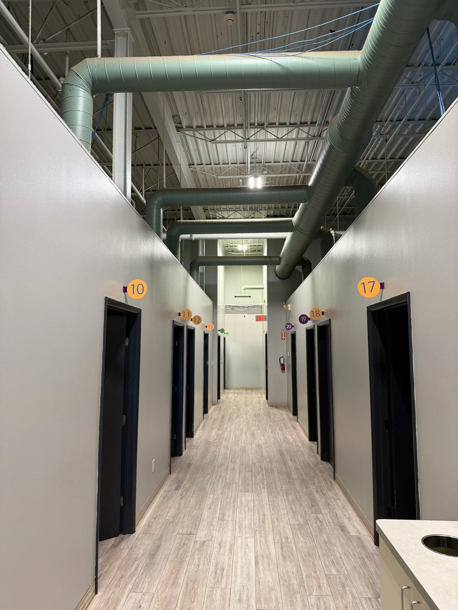 A hallway with small numbered private rooms on each side, featuring black doors and colorful number signs. Overhead industrial ceiling with visible ducts and pipes. The hallway has a wood-like vinyl floor and a fire extinguisher at the end.