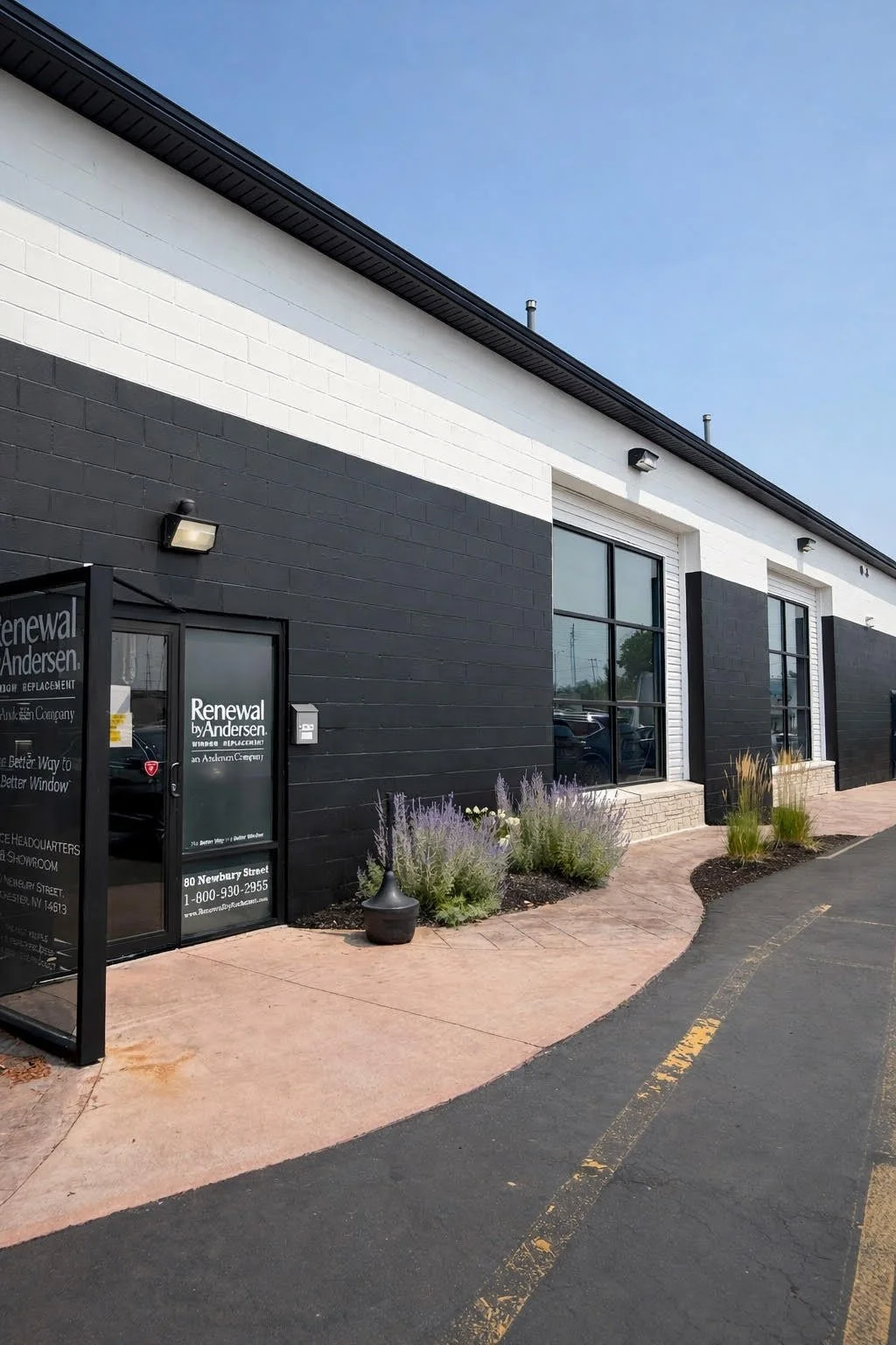 Exterior view of a commercial building with black and white painted brick walls, large windows, and landscaping with purple lavender plants. The building has business signage for 'Renewal by Andersen' near the entrance door.