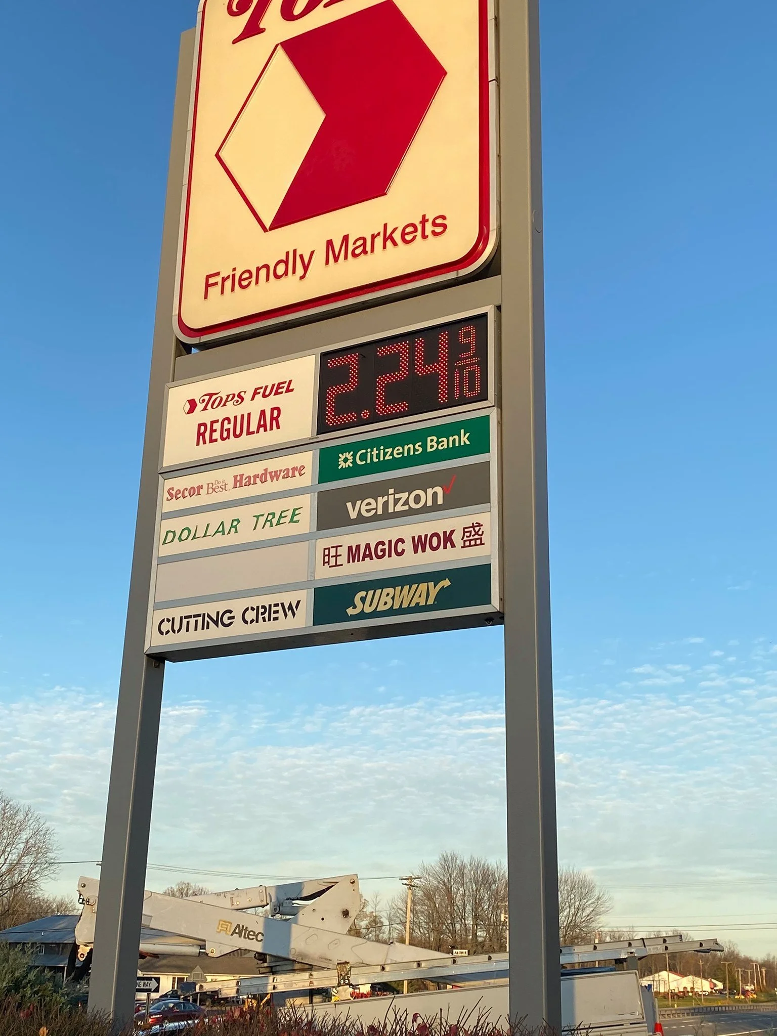 A sign at a gas station showing fuel prices and various business advertisements, with a blue sky in the background.