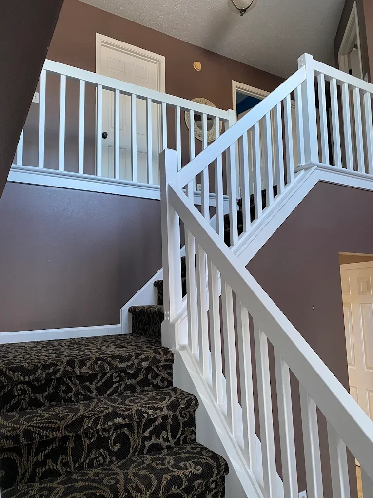 Indoor staircase with white railing and dark patterned carpet, leading to a second-floor landing with two closed white doors and wall decor, in a home interior.