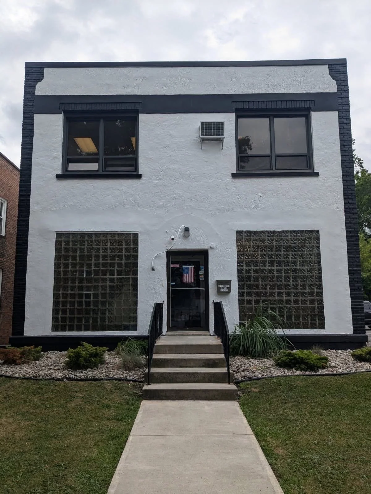 White two-story building with black accents and large glass block windows at ground level, a central door with steps and handrails, and two smaller windows on the upper level, with an air conditioning unit outside the upper right window.
