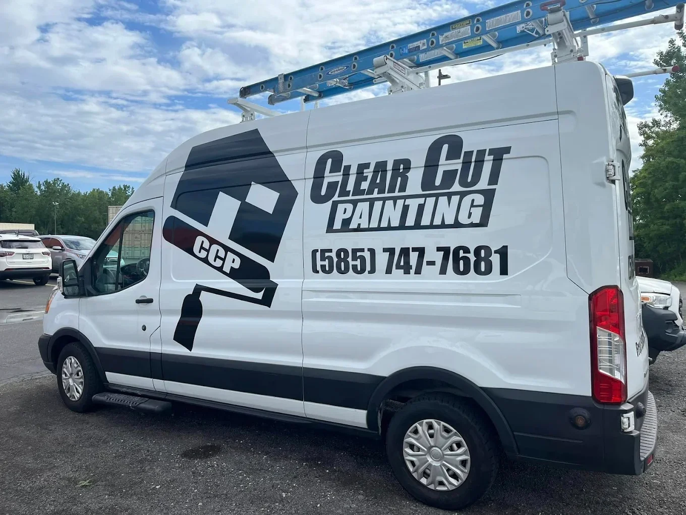 White service van with black and gray graphics advertising Clear Cut Painting, including a logo resembling a house with a paintbrush, phone number (585) 747-7681, parked in a lot with other vehicles, cloudy sky, and trees in the background.