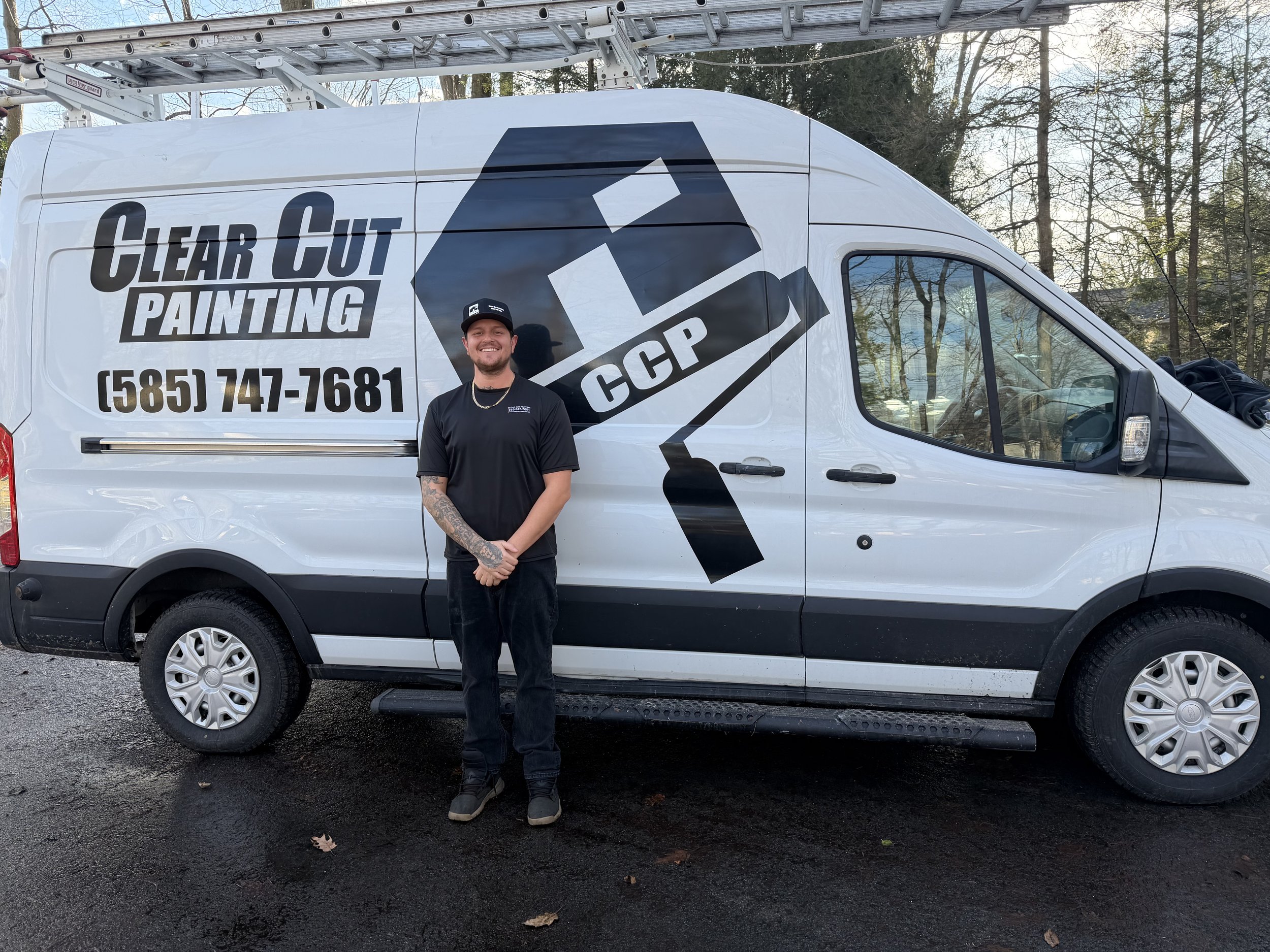 A smiling man with tattoos on his left arm standing in front of a white service van that has the text 'Clear Cut Painting' and a phone number, with a black and white logo of a paint roller and a paint can labeled 'CCP.' Trees with autumn foliage are in the background.