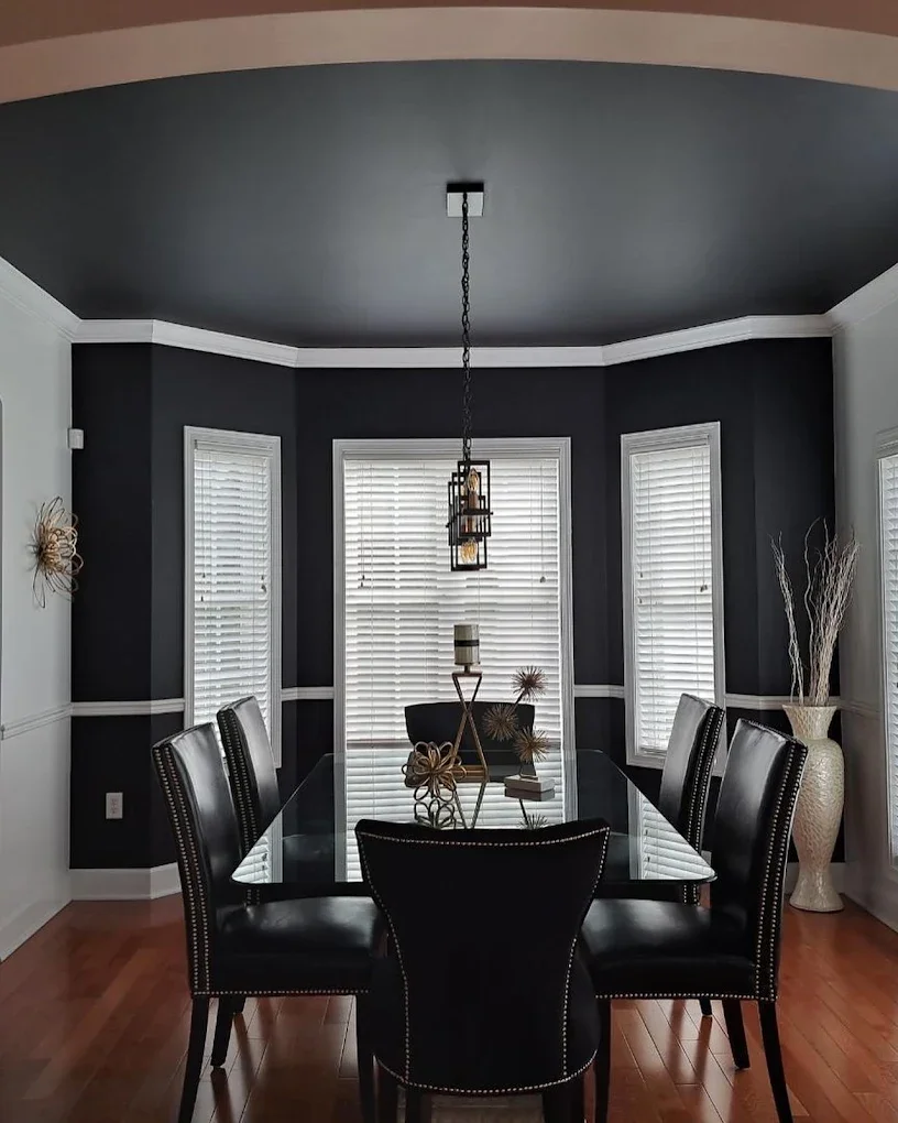 A modern dining room with a black glass table, six black leather chairs, and a chandelier hanging from a dark ceiling. The room has three windows with white blinds, white crown molding, and decor on the table. A large white vase with dried branches i