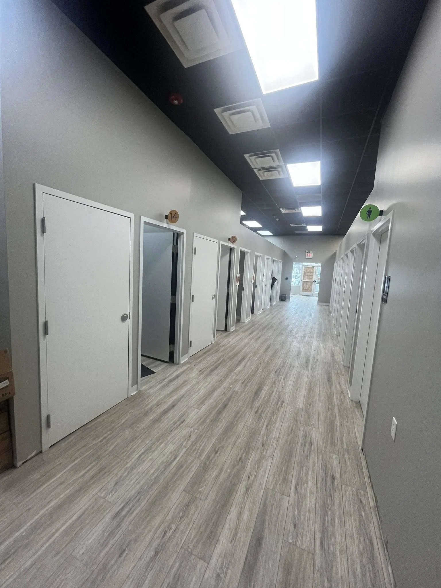 An indoor hallway with multiple small office or therapy rooms, each with a white door, numbered 12 through 14, on the left side, and a row of larger rooms with white doors on the right side. The flooring is wood-like, and the ceiling has black panels