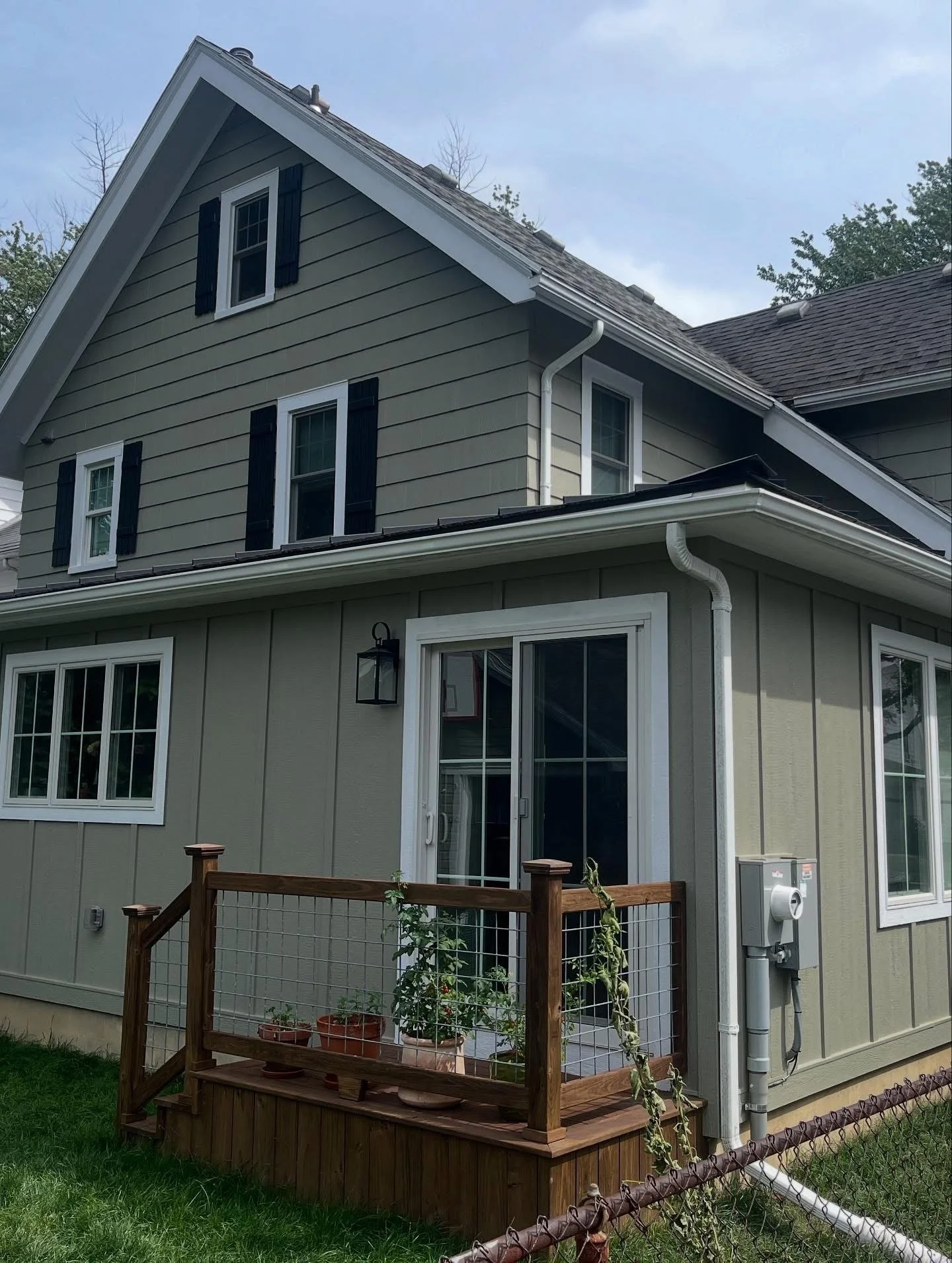 The image shows the exterior of a two-story house with beige siding and black shutters. There is a small wooden deck with potted plants and a glass sliding door. A black outdoor light fixture is mounted next to the door, and white gutters run along t