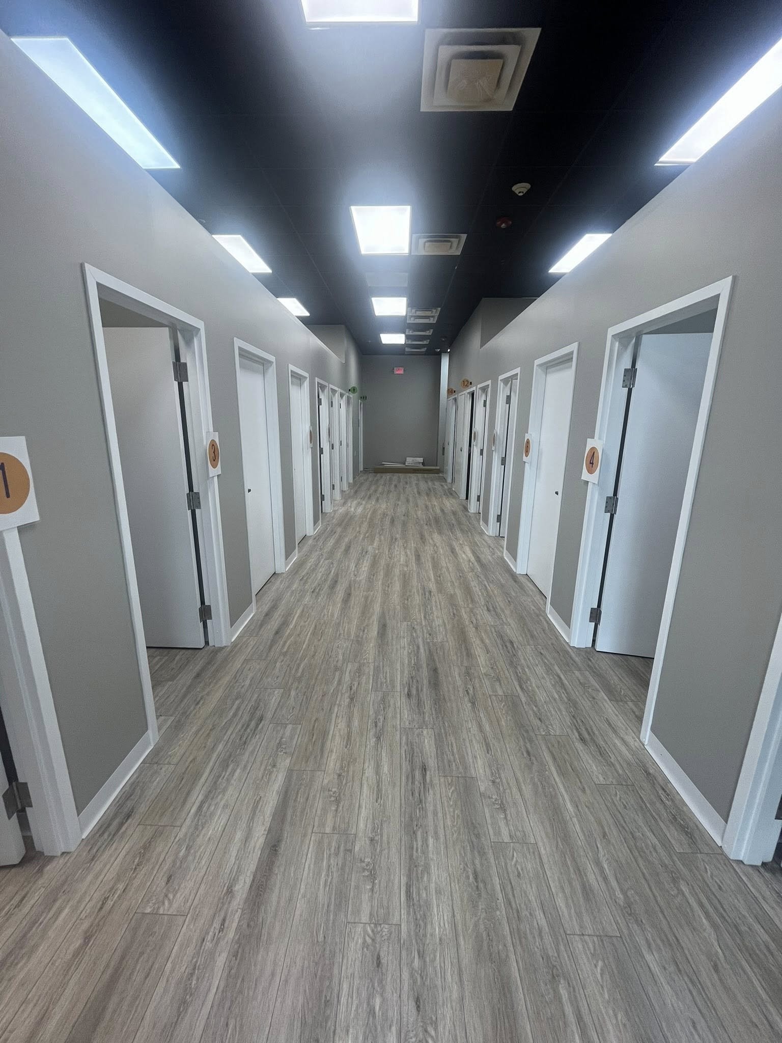 A hallway with multiple closed doors on both sides, numbered with round signs, and a wooden floor. The ceiling has bright rectangular lights and air vents.