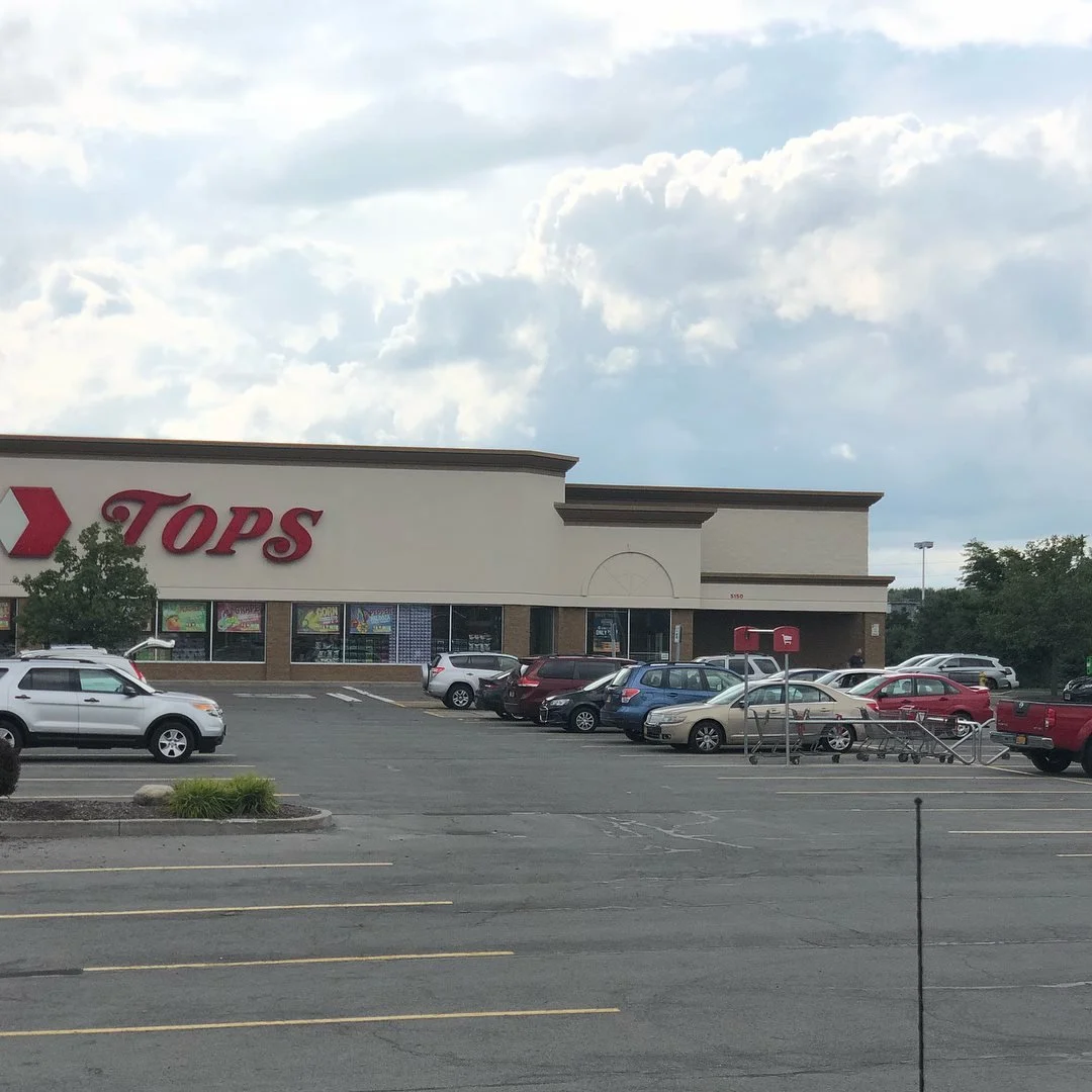 A shopping parking lot with several parked cars in front of a store called TOPS, which has a large red sign and shopping carts outside.