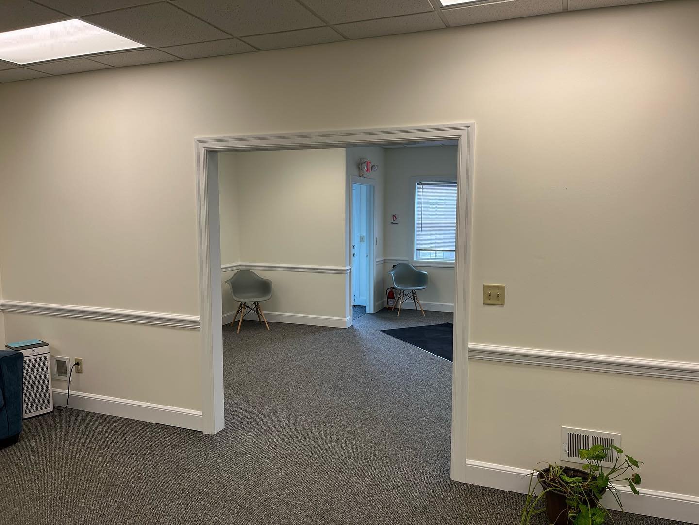 An office interior with beige walls and carpeted floors. There are two chairs, one in each room, near windows. A small indoor plant is on the right side near a wall vent. Part of a heater or air purifier is visible on the left side.