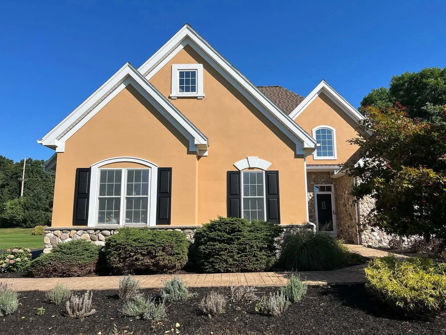 Front view of a two-story yellow house with white trim, black shutters, a stone foundation, and multiple gabled roofs, surrounded by landscaped bushes and plants under a clear blue sky.