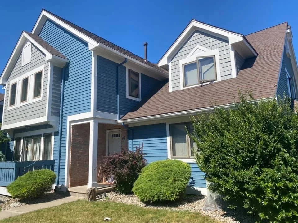 A two-story house with blue siding, white trim, a brown shingled roof, and several windows, surrounded by greenery and shrubs under a clear blue sky.