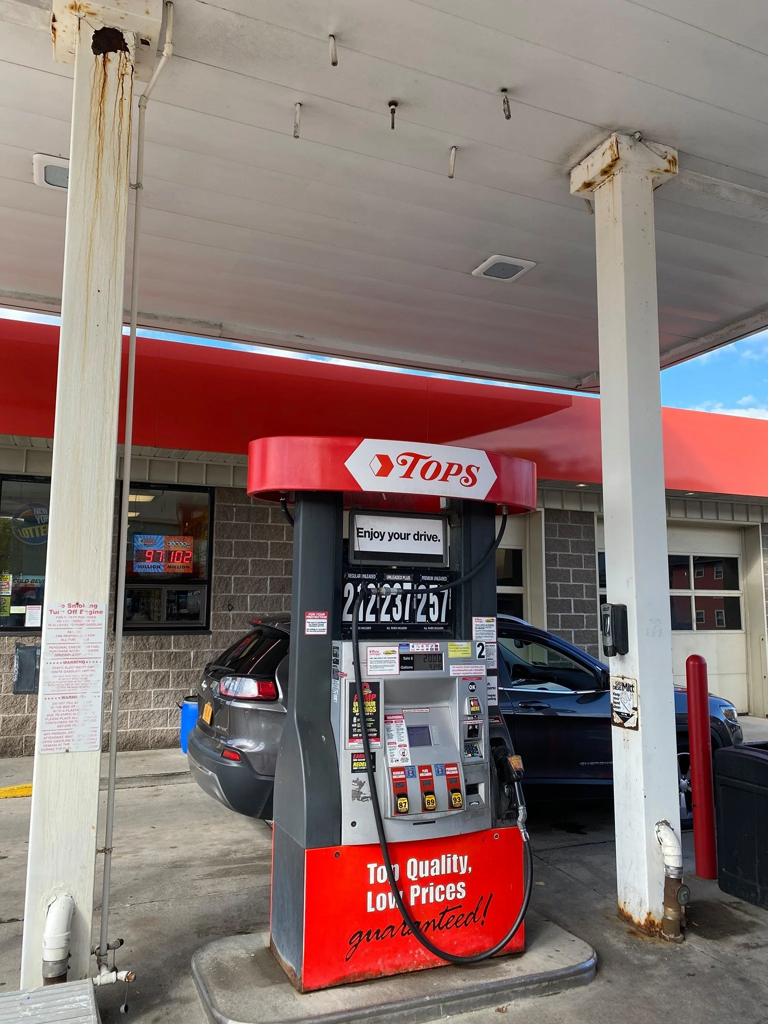 Gas pump at a gas station with a car parked underneath, and signs displaying gas prices, under a canopy.