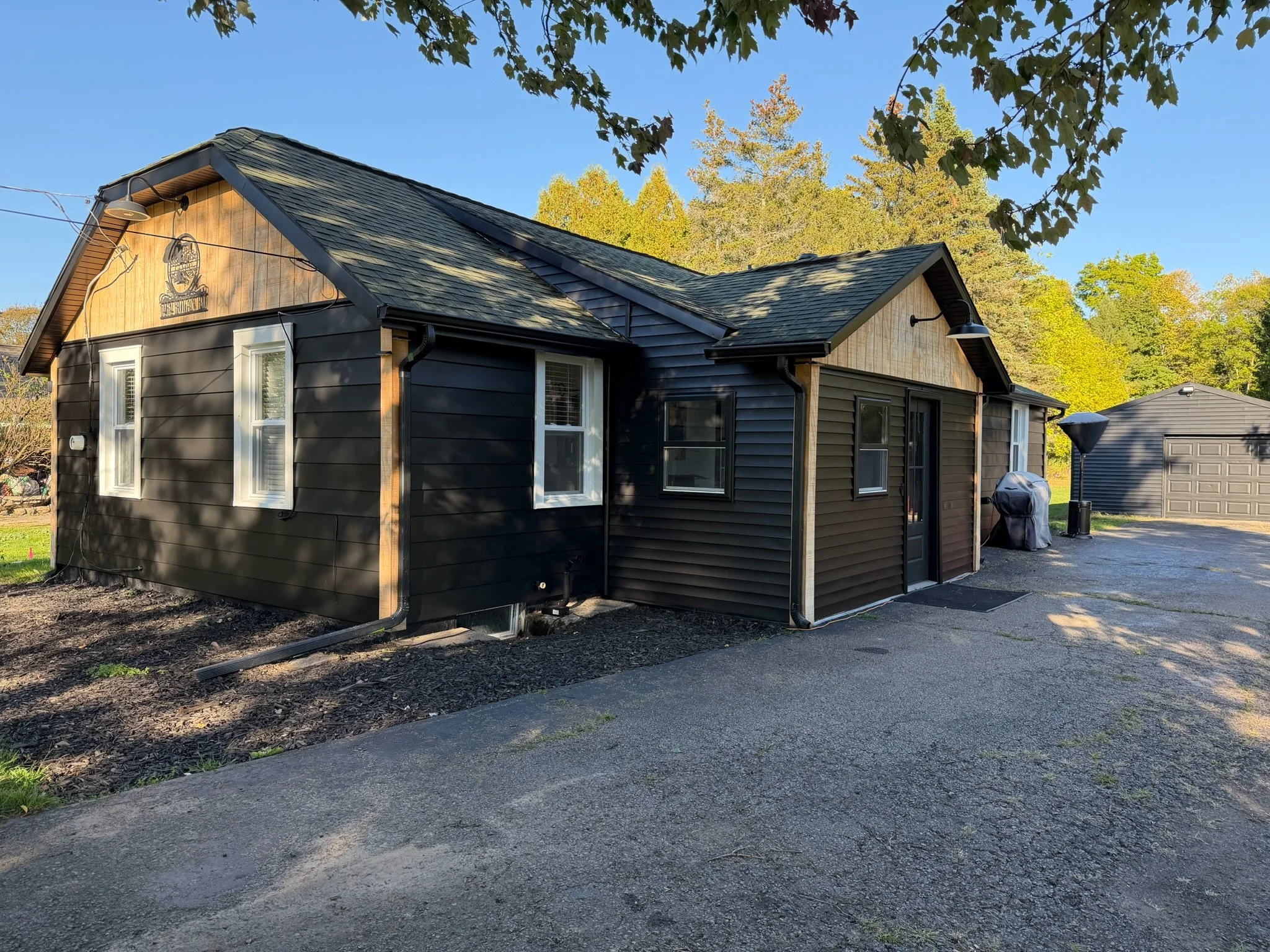 A small black house with a wood-paneled gable, white-framed windows, and a black door, situated next to a gravel driveway with a grill and outdoor light.