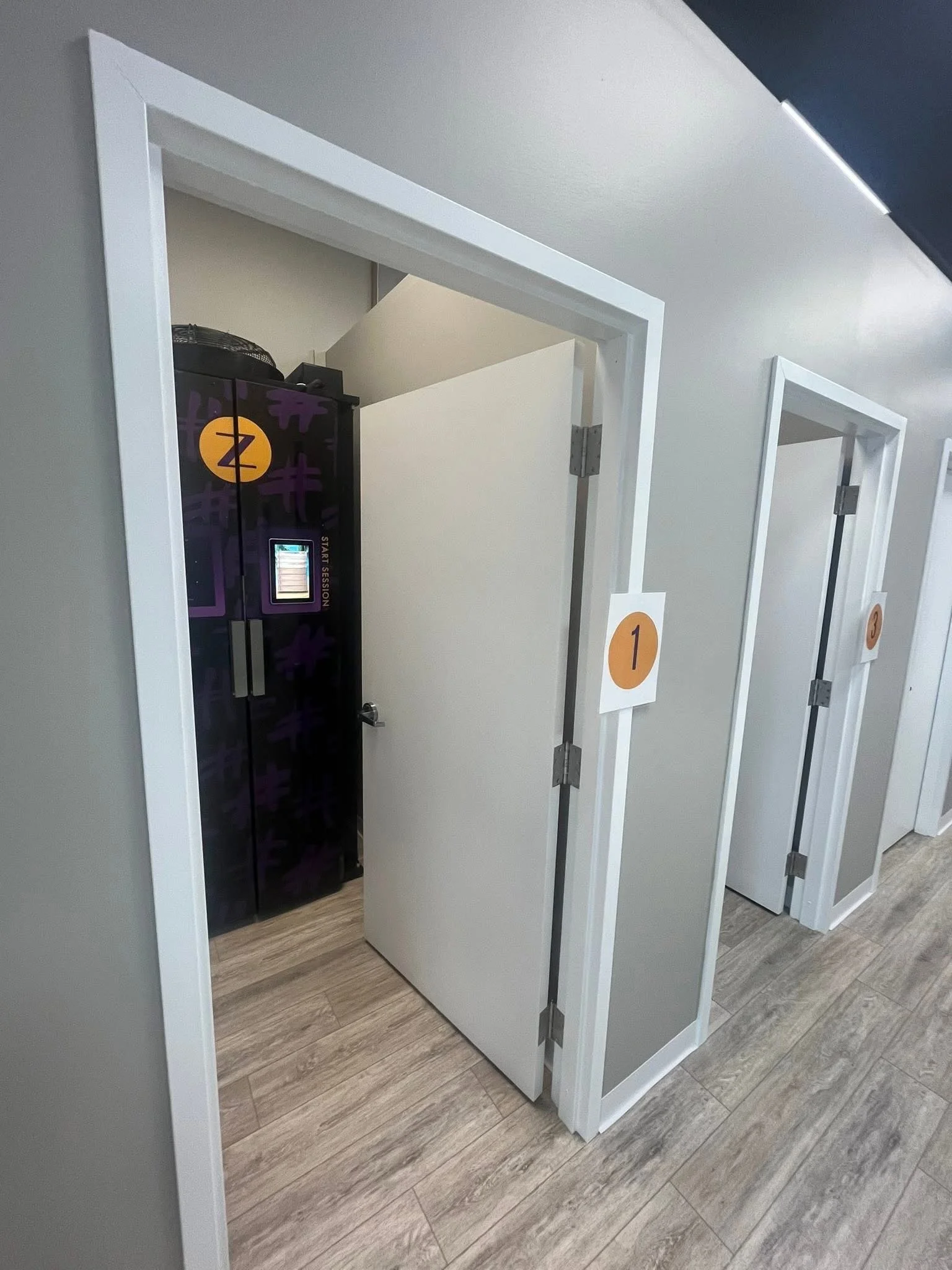 A row of three small rooms with open doors, numbered 1, 2, and 3, in an indoor setting with light-colored flooring and gray walls. Room 2 contains a black locker with purple graphics and a digital lock.