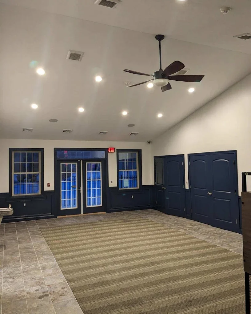 Empty room with tiled and carpeted flooring, white walls with dark blue wainscoting, and ceiling with multiple recessed lights. There are three windows and a double glass door with a window above, all framed in dark blue. A ceiling fan hangs from the
