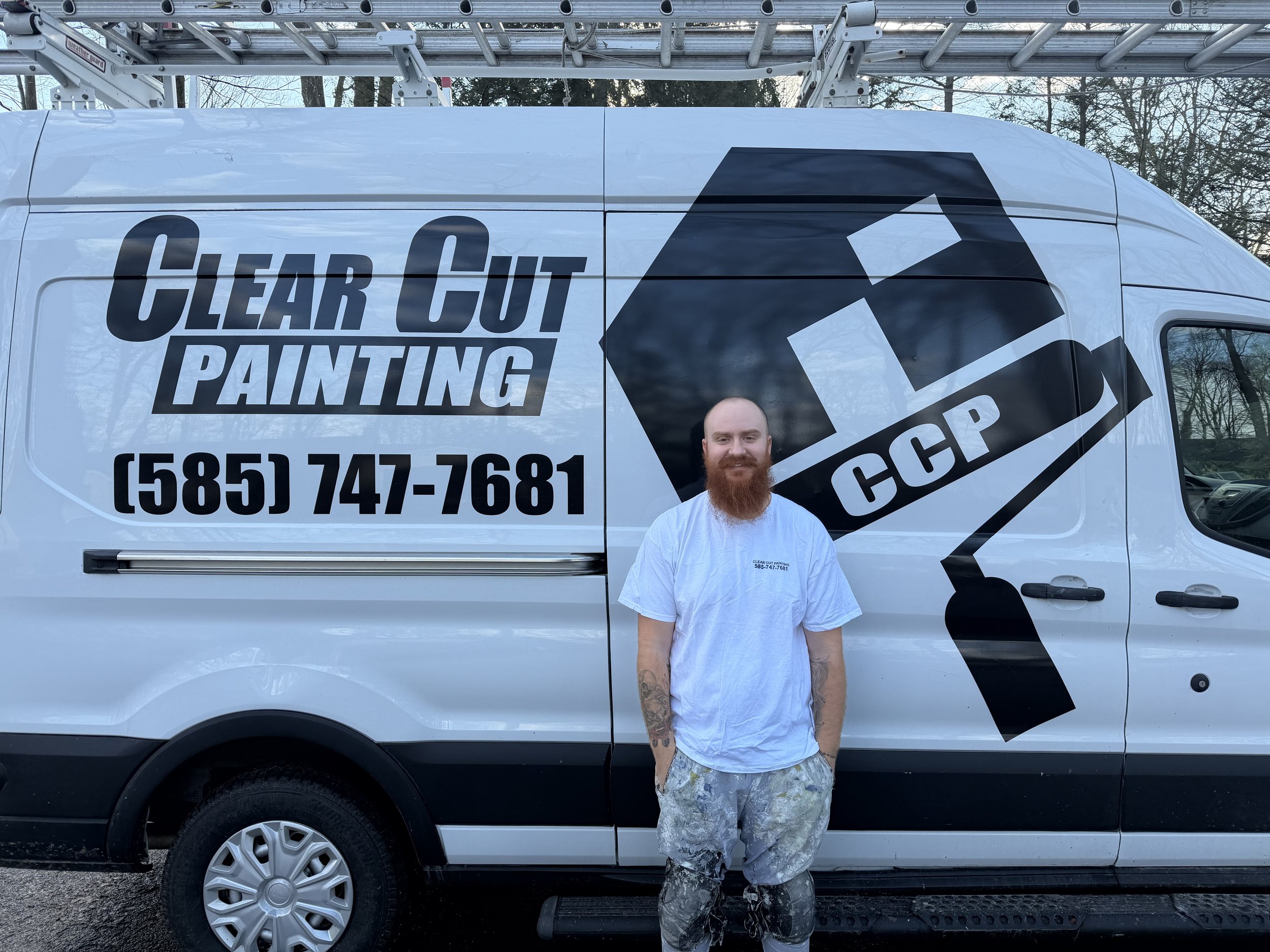 A man with a red beard and tattoos standing in front of a white work van with a large logo of a paint roller and the text "Clear Cut Painting" and a phone number. The man is wearing a white T-shirt and paint-splattered pants.