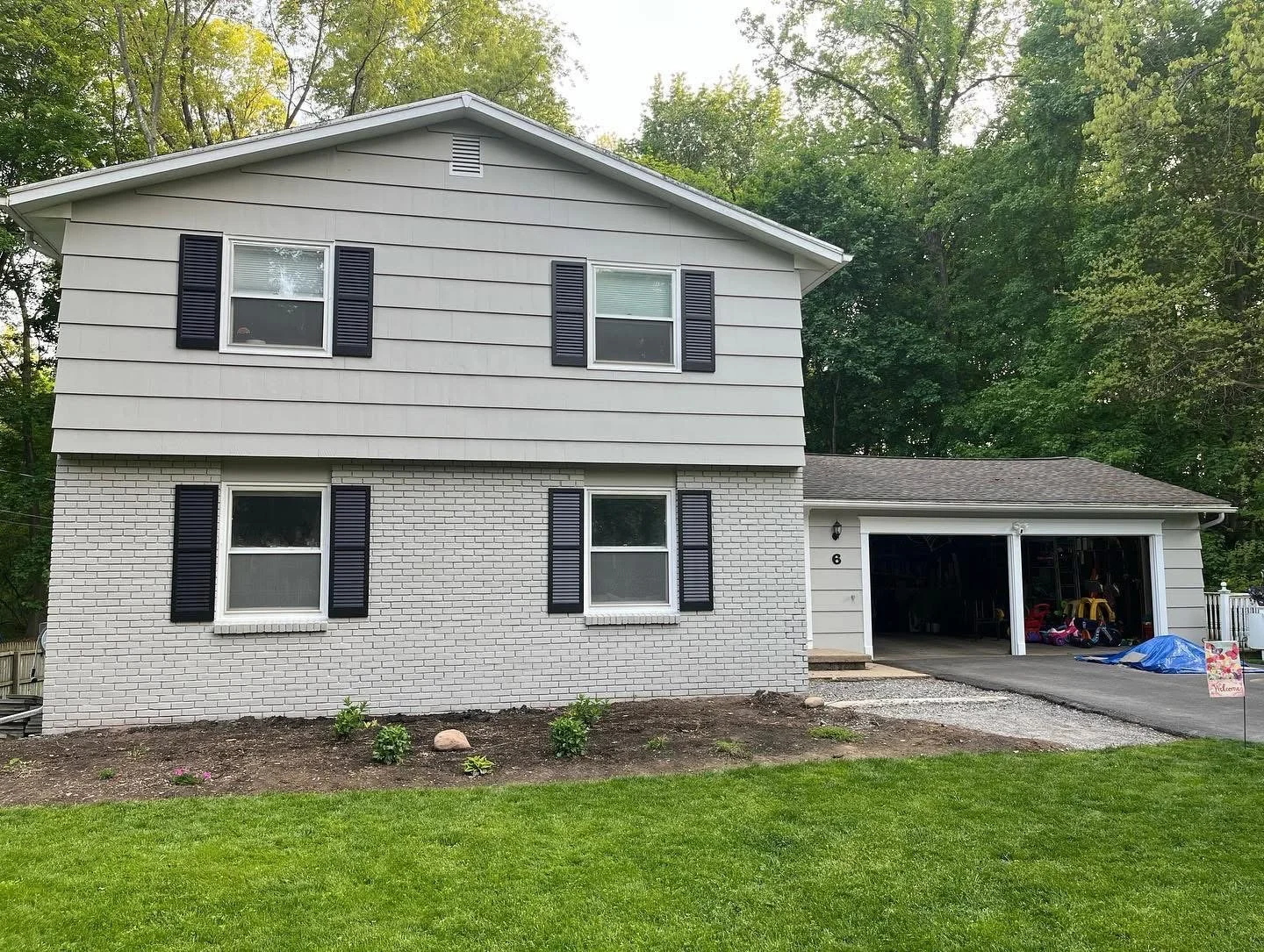 A two-story house with gray siding on the upper level and white brick on the lower level, black shutters on the windows, and an attached garage with some stored items visible inside. The house is bordered by a patch of grass and under construction gr