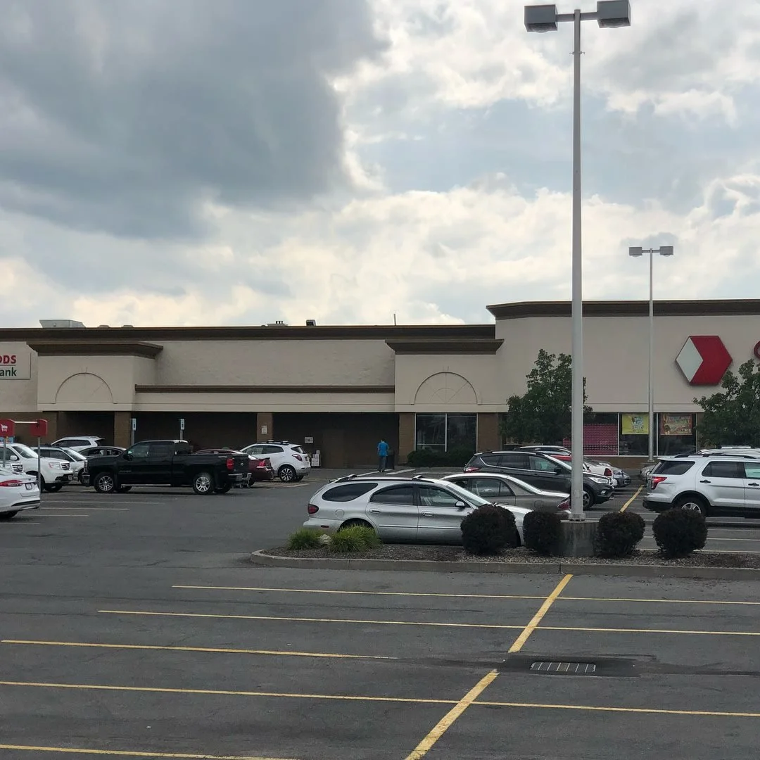 View of a shopping plaza parking lot with several parked cars, some trees, and a building with a red logo. The sky is cloudy with some sunlight visible.