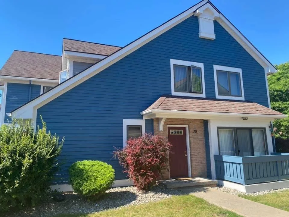 A blue two-story house with white trim, a small front porch, and shrubs in the yard under a clear blue sky.
