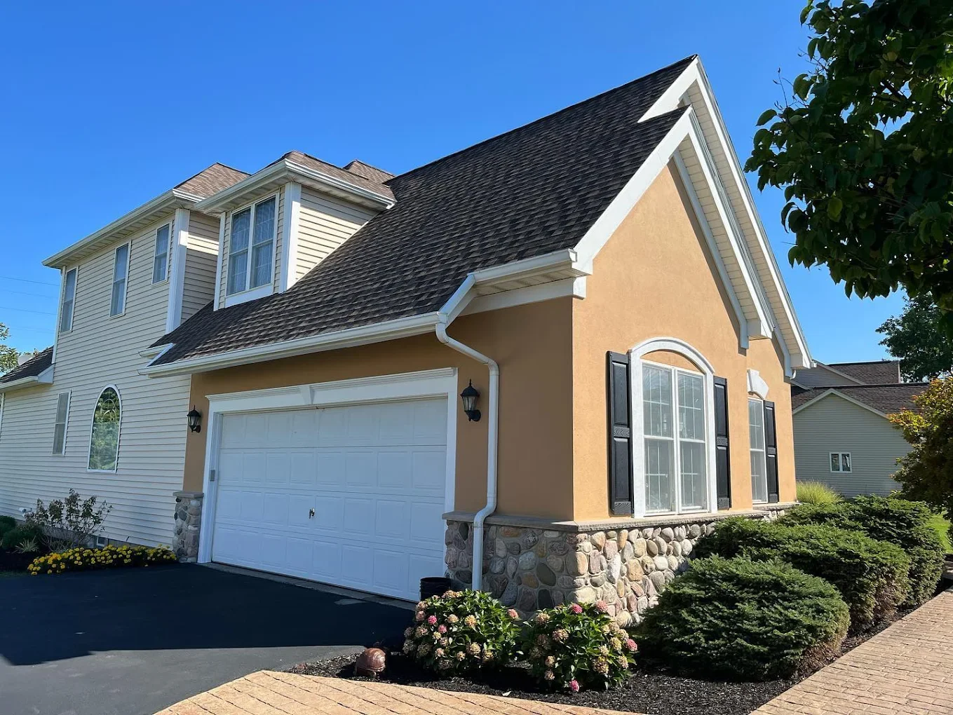 Front view of a house with a yellow facade, large arched window with black shutters, stone foundation, and a white garage door. The house has a steep roof with brown shingles, white trim, and is surrounded by landscaped bushes, flowers, and a brick w