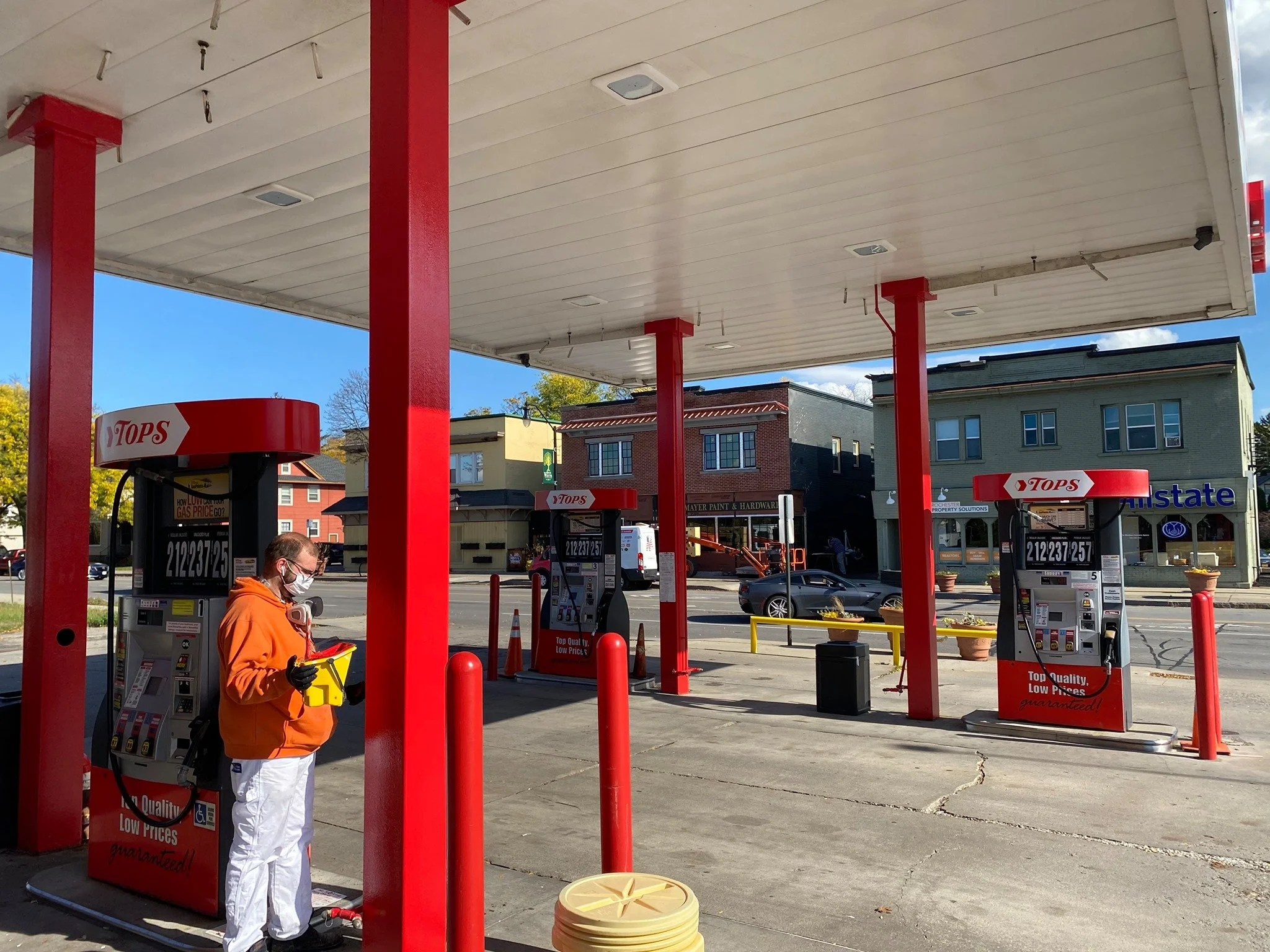 A gas station with three pumps, a person wearing an orange hoodie, white pants, a mask, and gloves, standing near a pump, holding a yellow payment terminal, in a small town with buildings across the street and a clear sky.