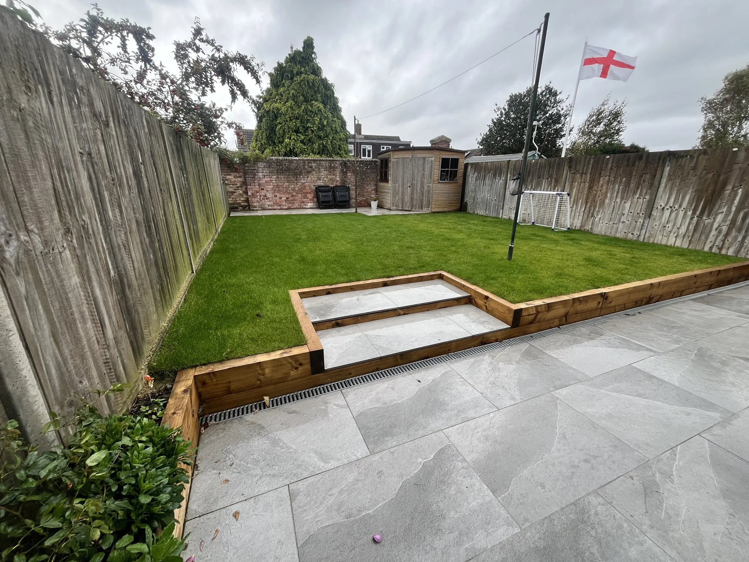 Backyard with a grassy lawn, a small wooden shed, a flagpole with a flag, and a soccer goal, surrounded by wooden fencing and a paved patio area.
