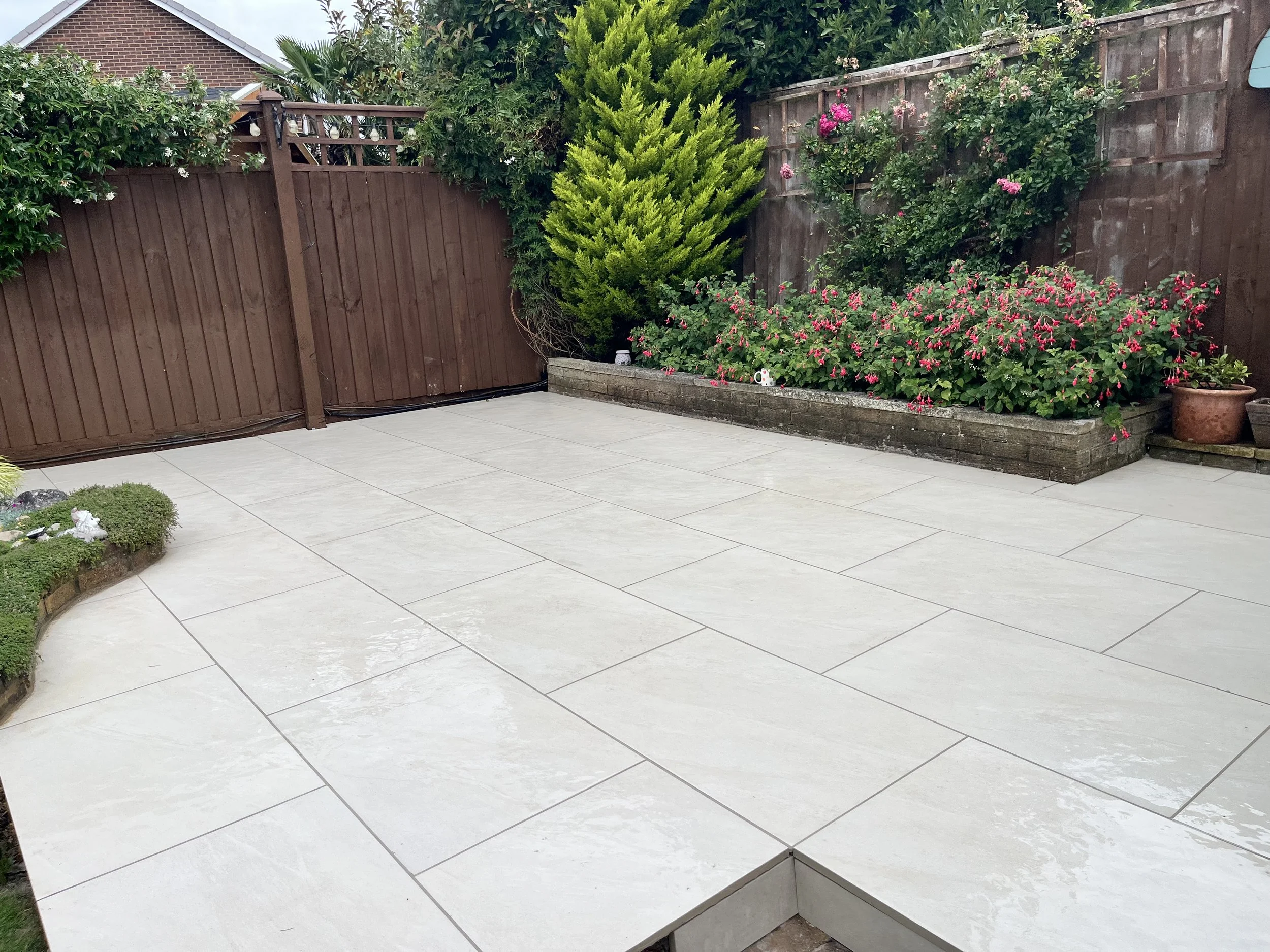 Backyard patio with large beige stone tiles, bordered by a red wooden fence, with lush green plants, shrubs, and pink flowering bushes along the fence line.