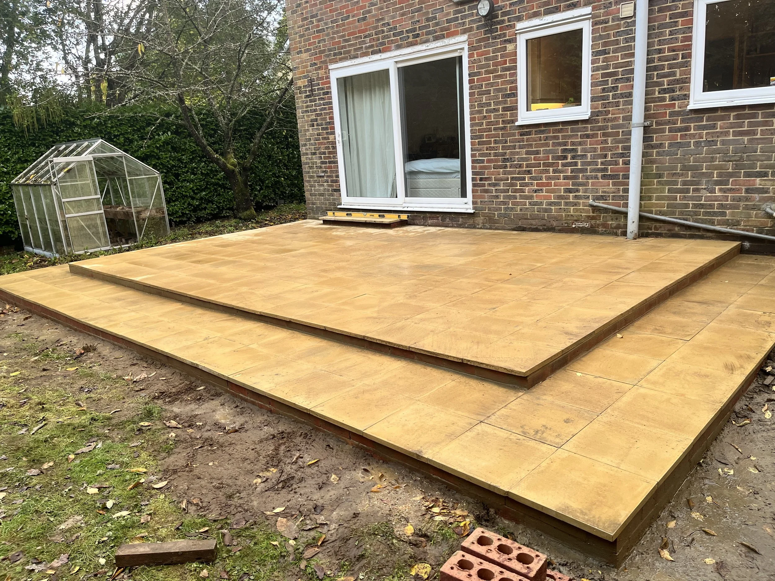 Newly installed wooden patio with two levels in a backyard next to a brick house, with sliding glass door, windows, a small greenhouse, and a tree with bare branches in the background.