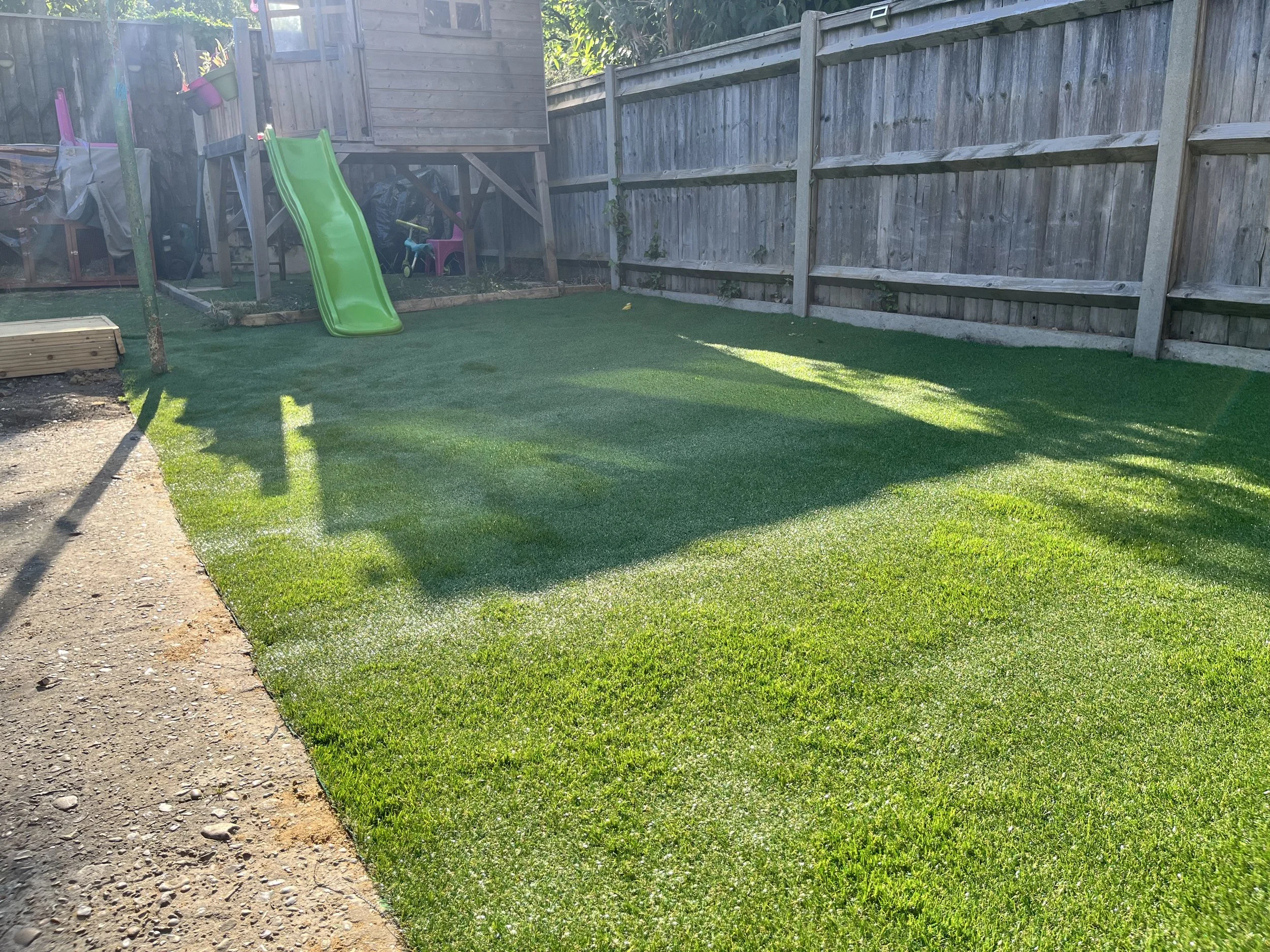Well-maintained backyard with a small green slide, a wooden playhouse, and a fenced perimeter, featuring a mix of grass and gravel surfaces.