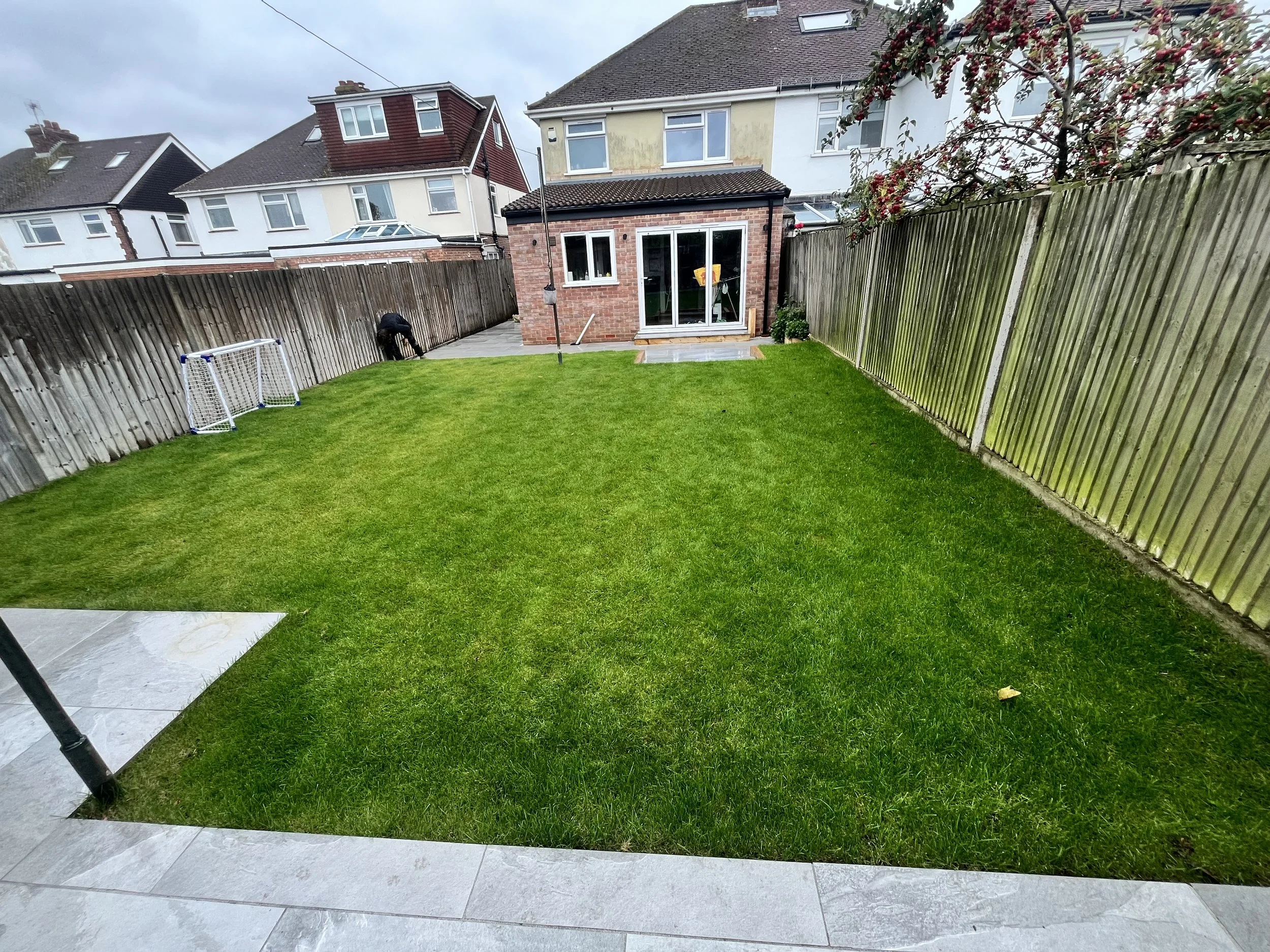 View of a backyard with a green lawn, a wooden fence on the right, and concrete patio in the foreground. There are two small soccer goals on the left, a clothesline hanging in the middle, and a person near the fence on the far left. The house has lar