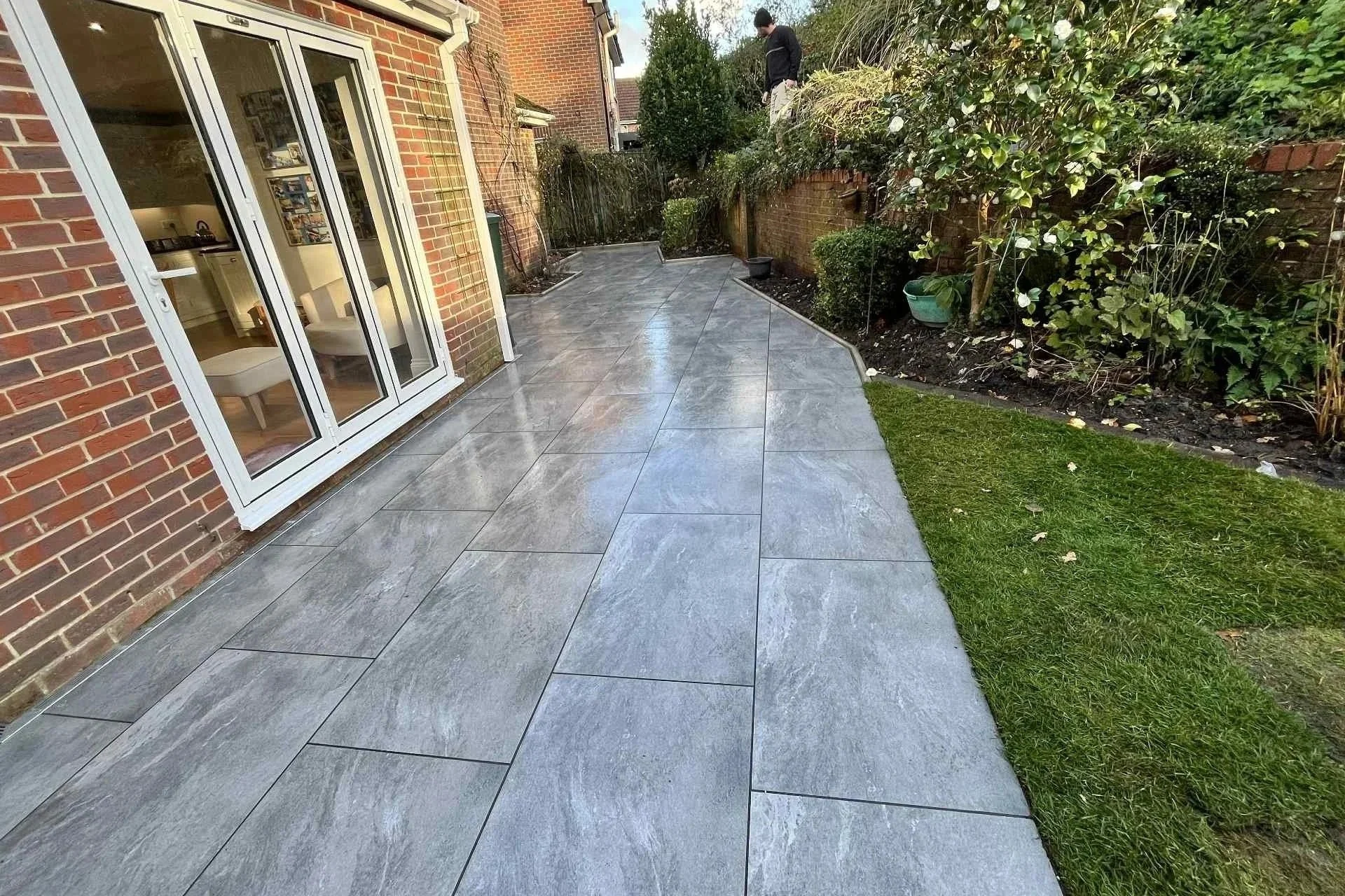 A newly tarmacked patio with large gray tiles next to a brick house with white framed glass doors. There are green plants and bushes along the garden border, with a man standing at the far end of the garden near a small tree.