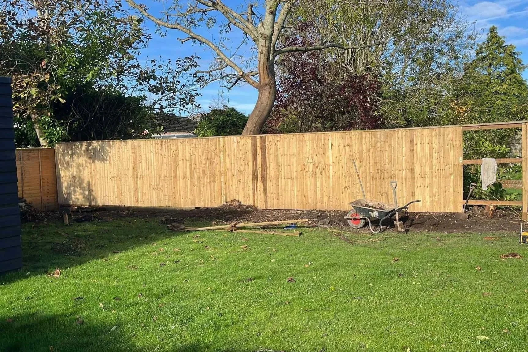 backyard with a newly installed wooden fence, construction tools, and a wheelbarrow, trees in the background, sunny day