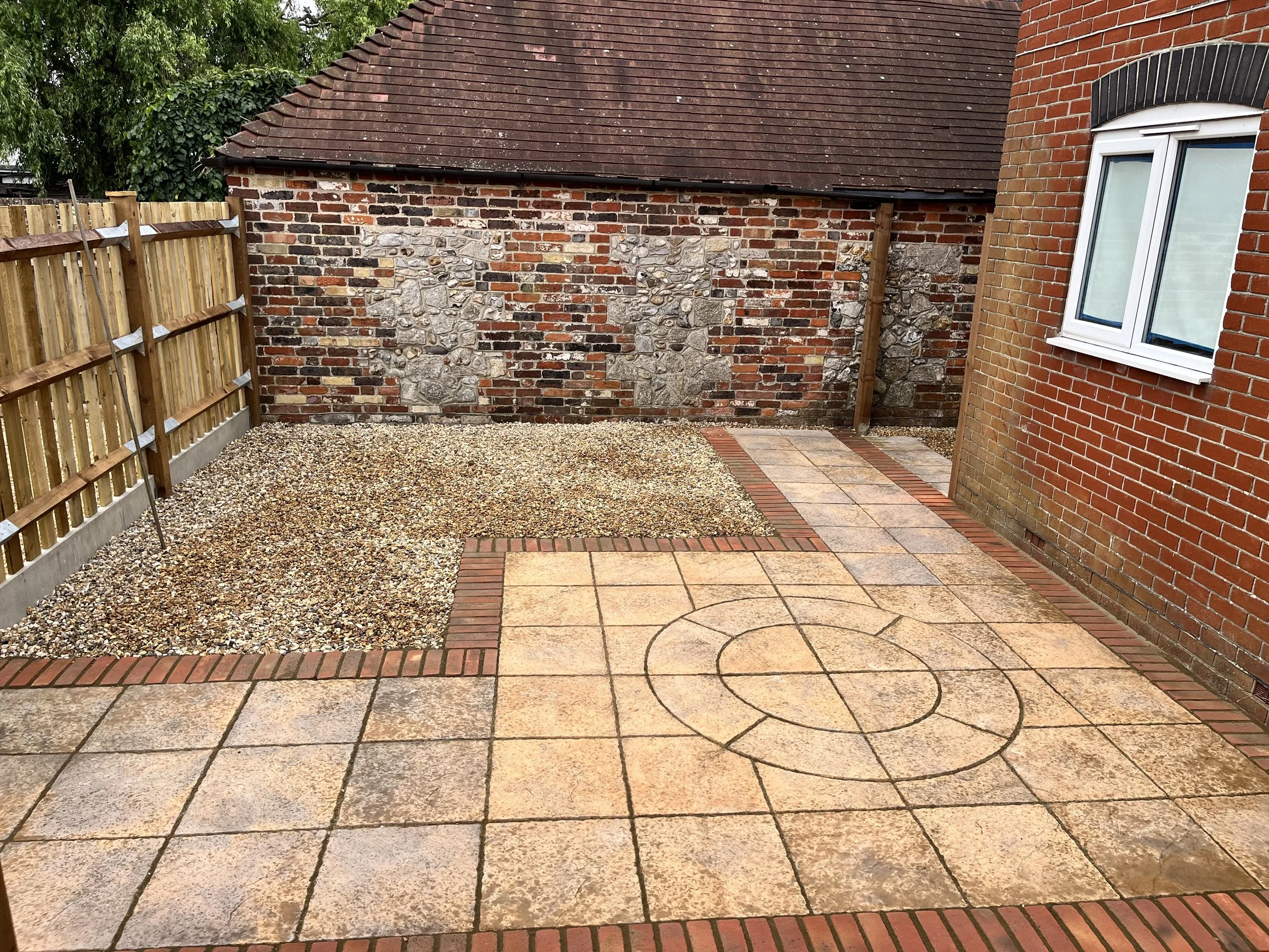 A patio with large beige paving stones featuring a circular pattern, bordered by red bricks. To the left, there is a gravel area enclosed by a wooden fence. The background includes a brick wall with stone patches and a neighboring brick building with