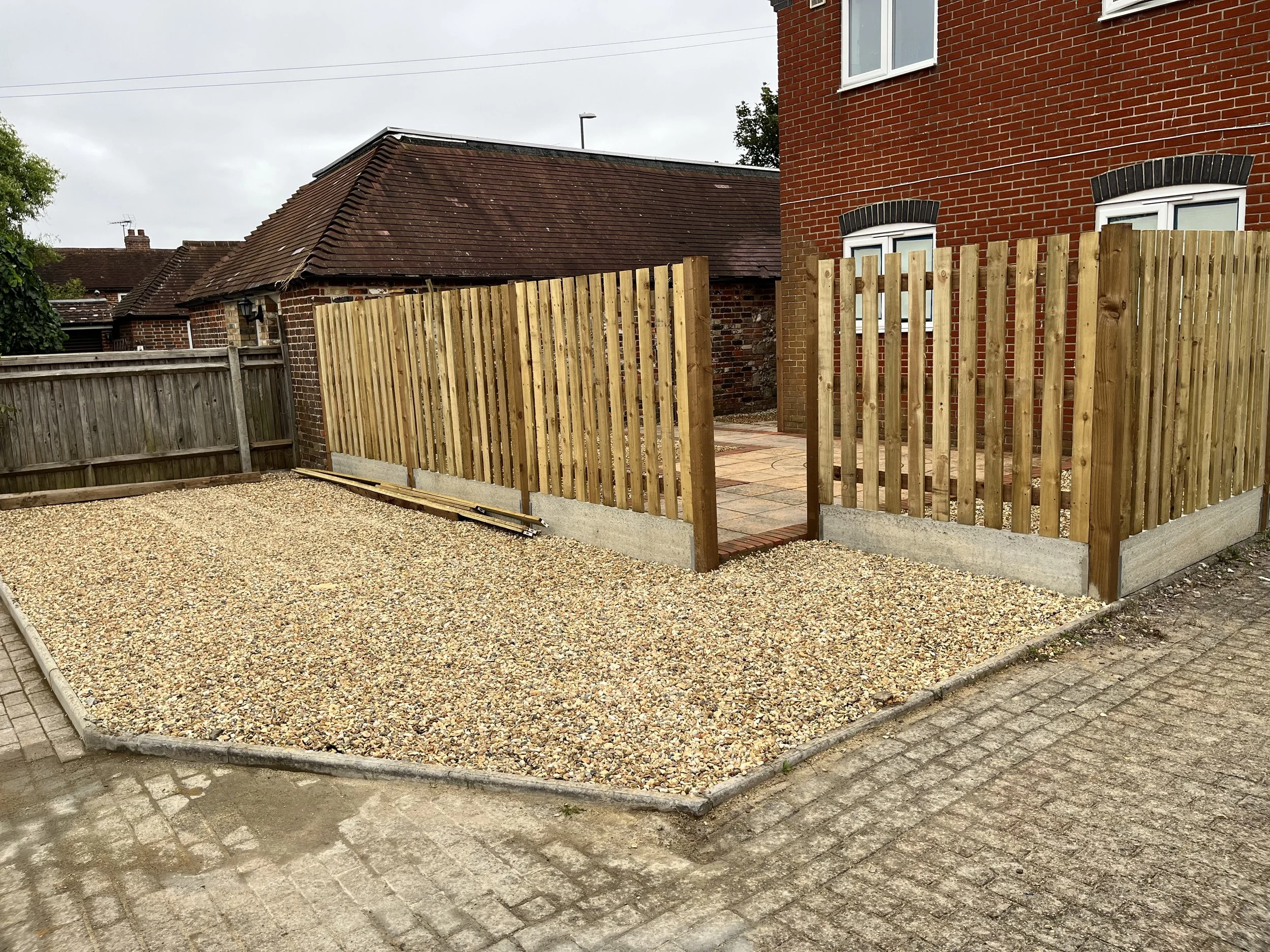 Partially constructed wooden fence around a backyard with gravel and paving stones in front of a brick house.