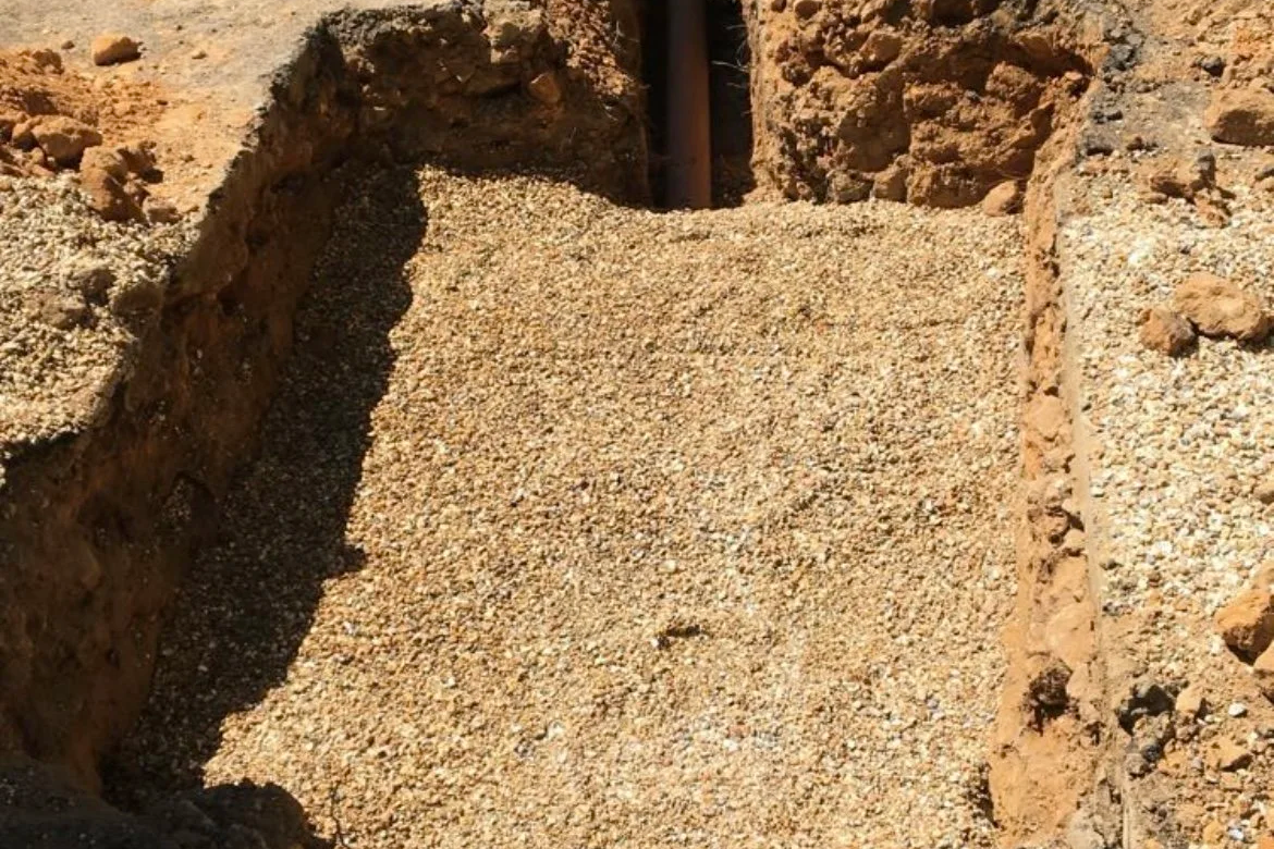 A trench filled with small rocks and gravel, with excavated dirt on both sides and a pipe visible at the top.