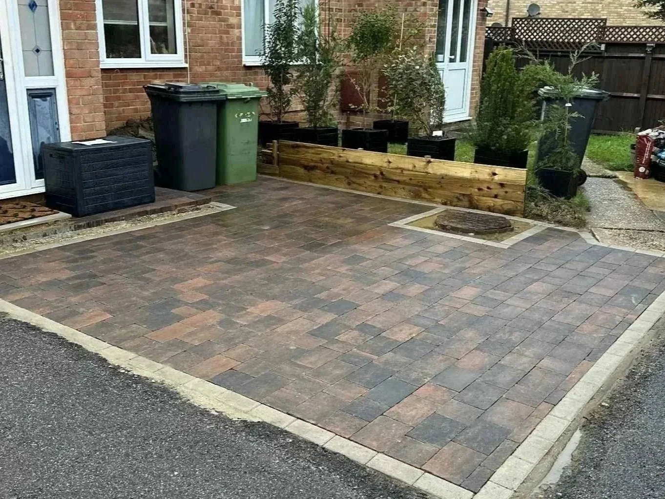 A newly paved small driveway or patio area with interlocking bricks next to a brick house. There are two trash bins, one black and one green, near the house. A wooden garden bed with small trees and potted plants is along the back edge. Gravel pathways lead to the side of the house, and in the background, there is a wooden fence.