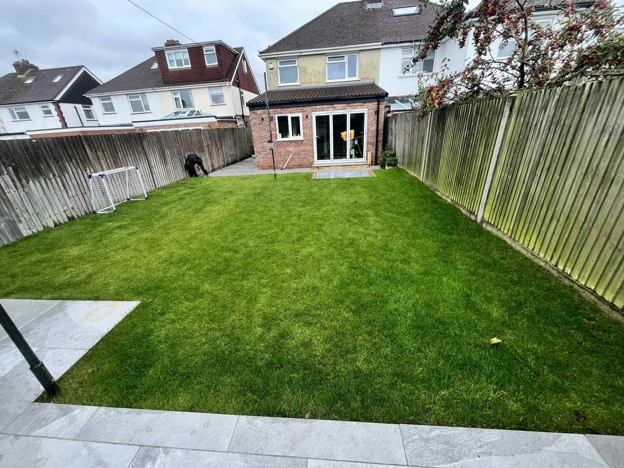 Garden with a grassy lawn, wooden fence on both sides, a small soccer goal, a patio area, and a brick house with white sliding glass doors. Overcast sky.