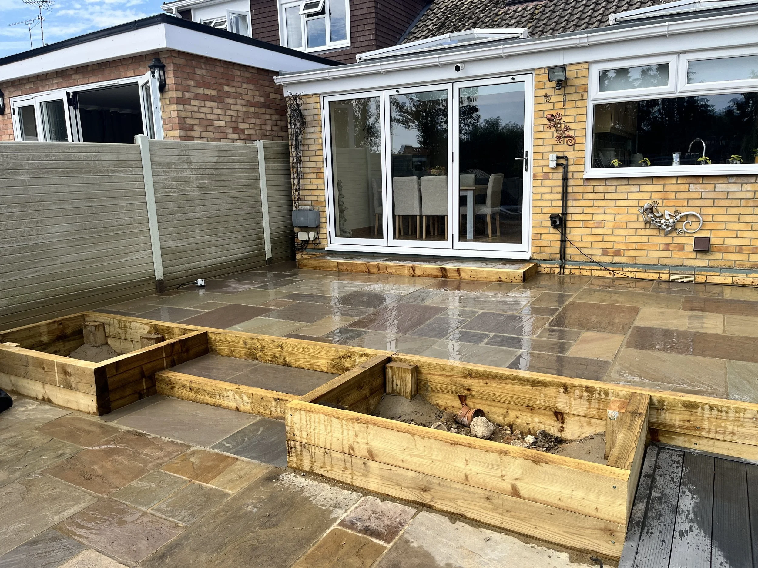 Backyard patio with stone flooring, partially built wooden planters, a patio table with chairs inside a screened-in area, and a brick house with decorative metal lizard and frog wall hangings.
