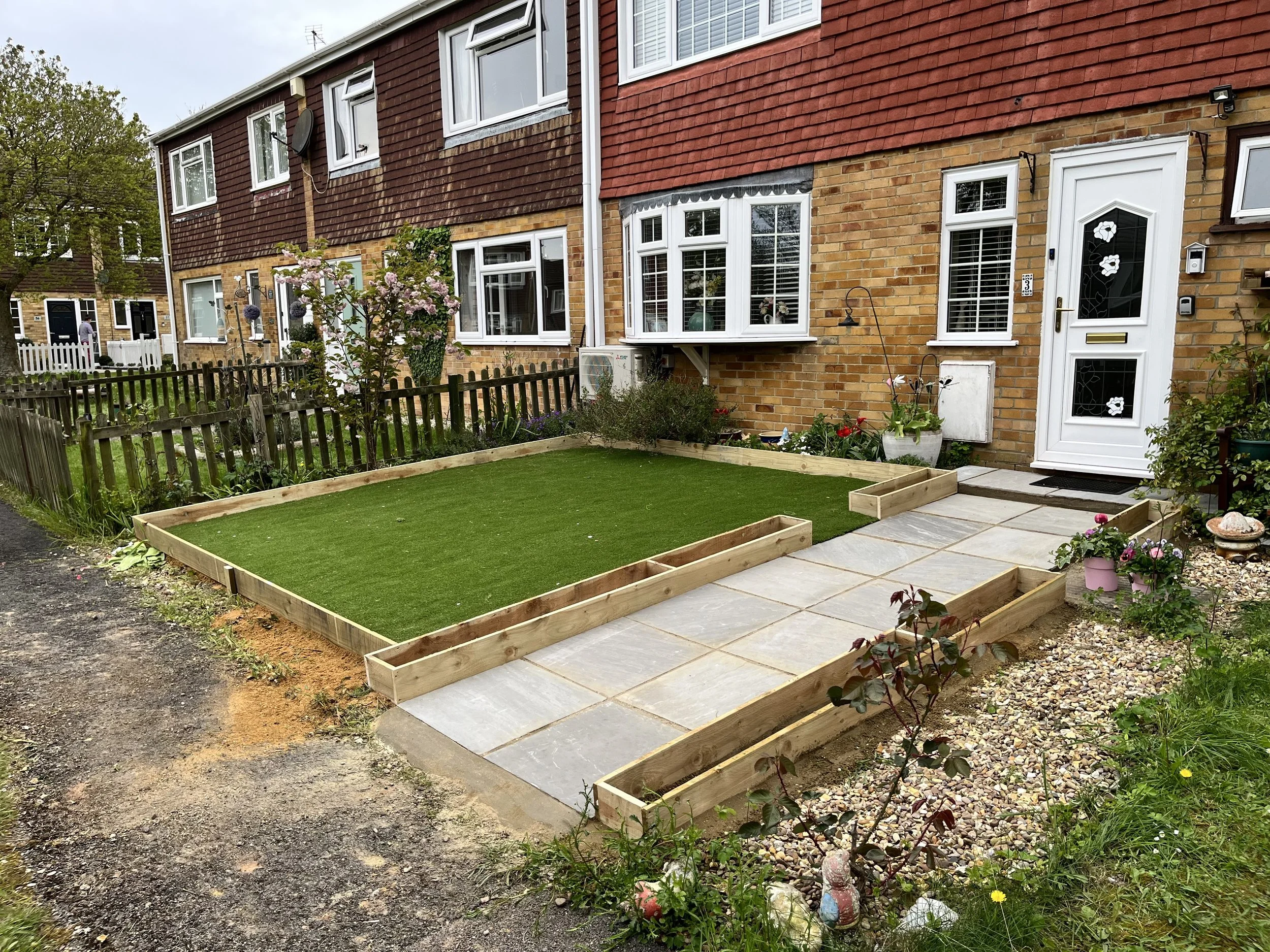Front yard of a brick house with a partially built small garden with wooden borders and artificial grass, paving stones leading to the door, and various flowering plants.