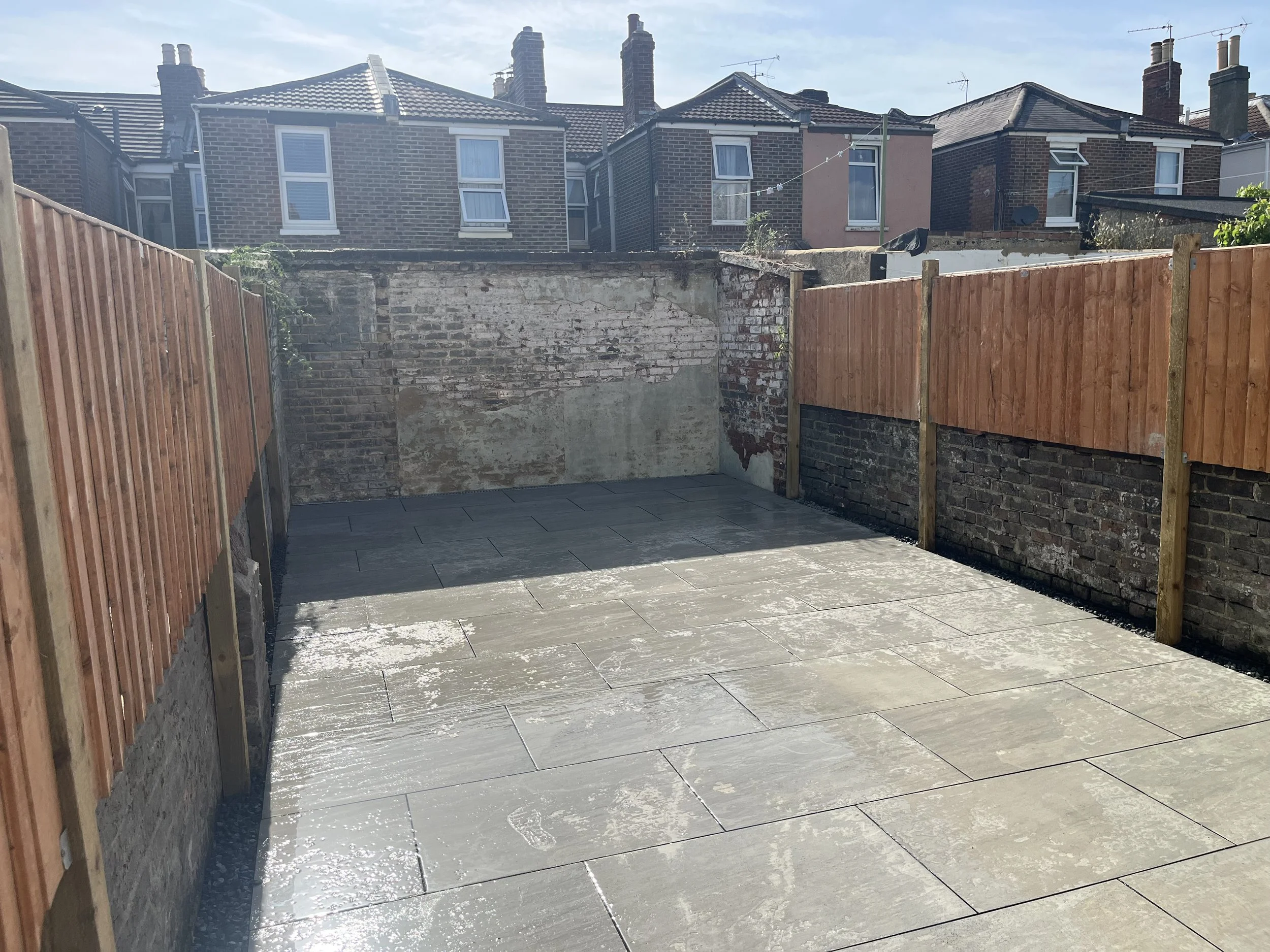 Empty backyard with newly installed grey stone pavers, brick and concrete wall at the back, and wooden fences on both sides, under a clear blue sky.