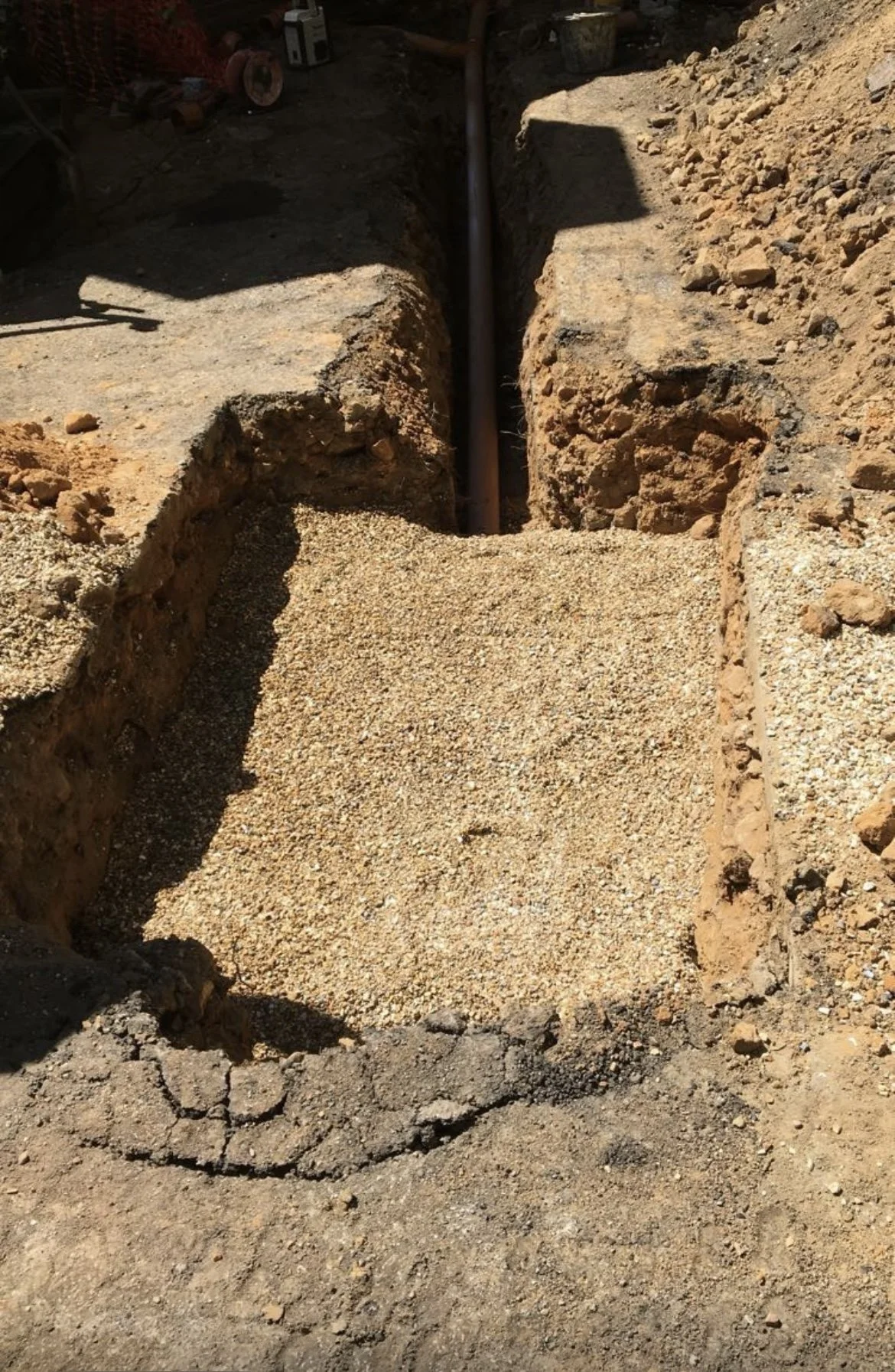 A trench with a pipe at the bottom, surrounded by dirt and gravel, in a construction site.