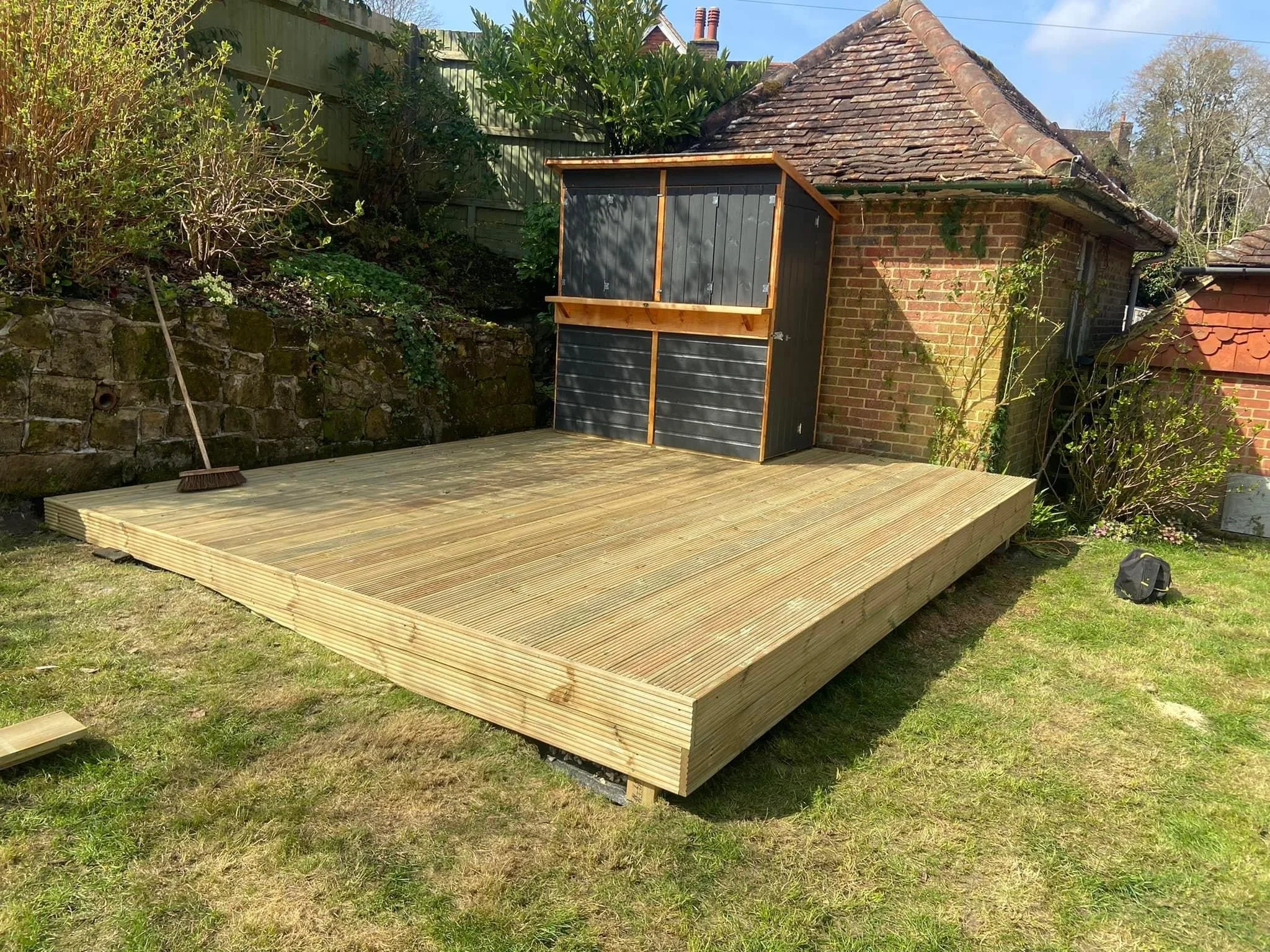 A small wooden deck in a backyard next to a brick shed with a black and wood storage unit on its side. A broom is leaning against a stone wall to the left, and a black bag is on the grass to the right. The background includes trees and a blue sky.