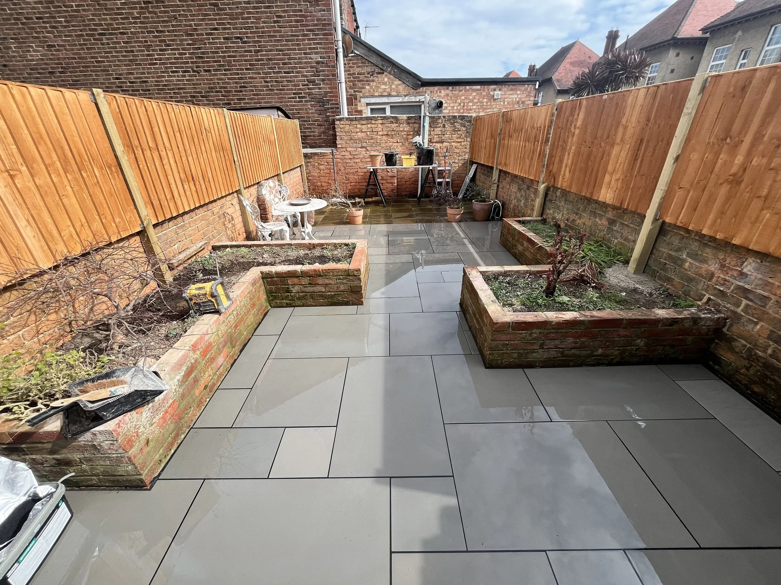 Backyard with new wooden fence panels, potted plants, garden tools, and a small white metal table and chairs on tiled ground.