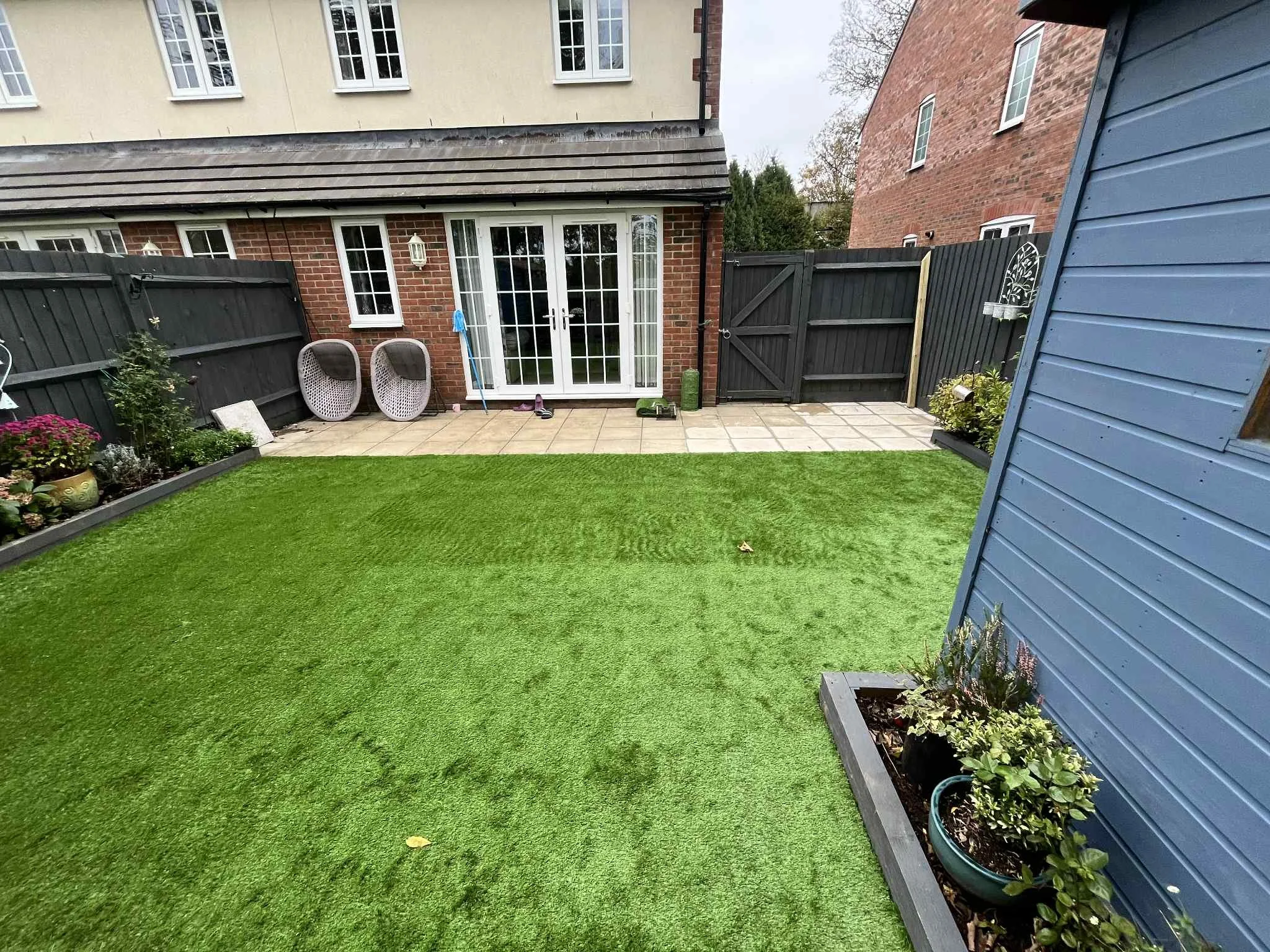 A garden with a green lawn, potted plants, a patio area with outdoor chairs, and sliding glass doors leading into a house, enclosed by a black wooden fence.