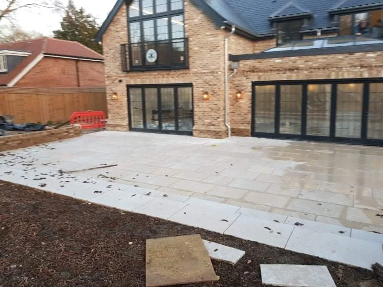 Backyard patio under construction with paving stones, adjacent to a brick house with large glass doors and windows. There is a wooden fence and some construction materials on the side.