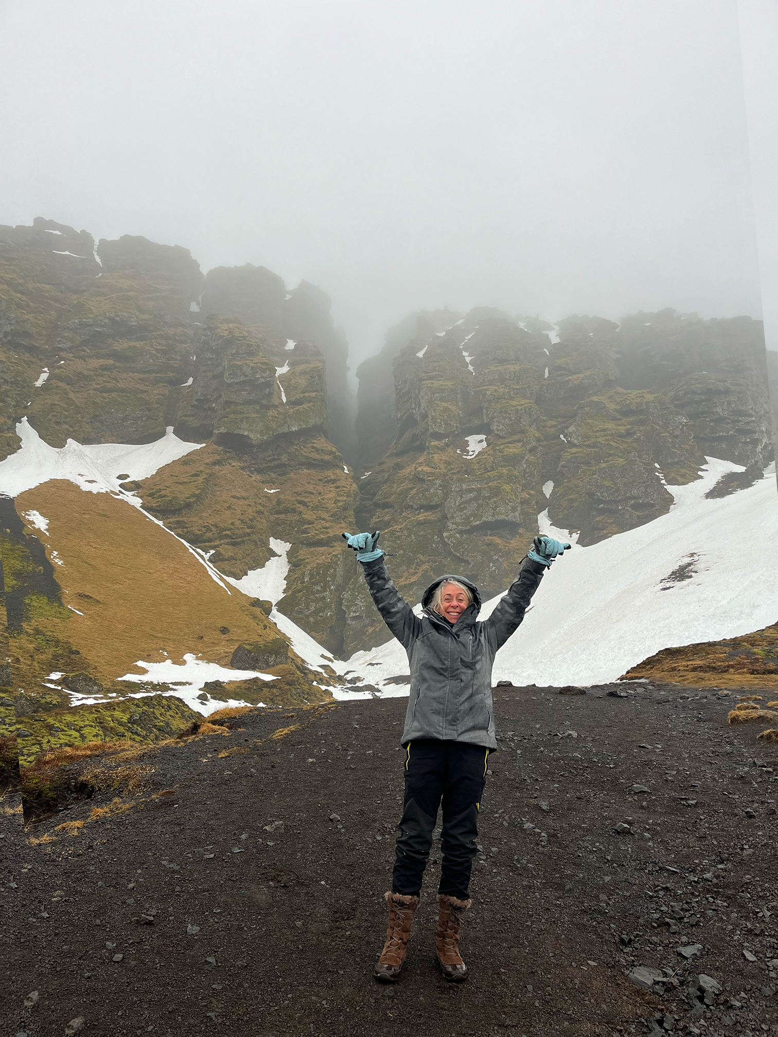 A woman dressed in outdoor hiking gear with her arms raised celebrating on a volcanic rocky terrain with snow patches, misty mountains, and foggy sky in the background.