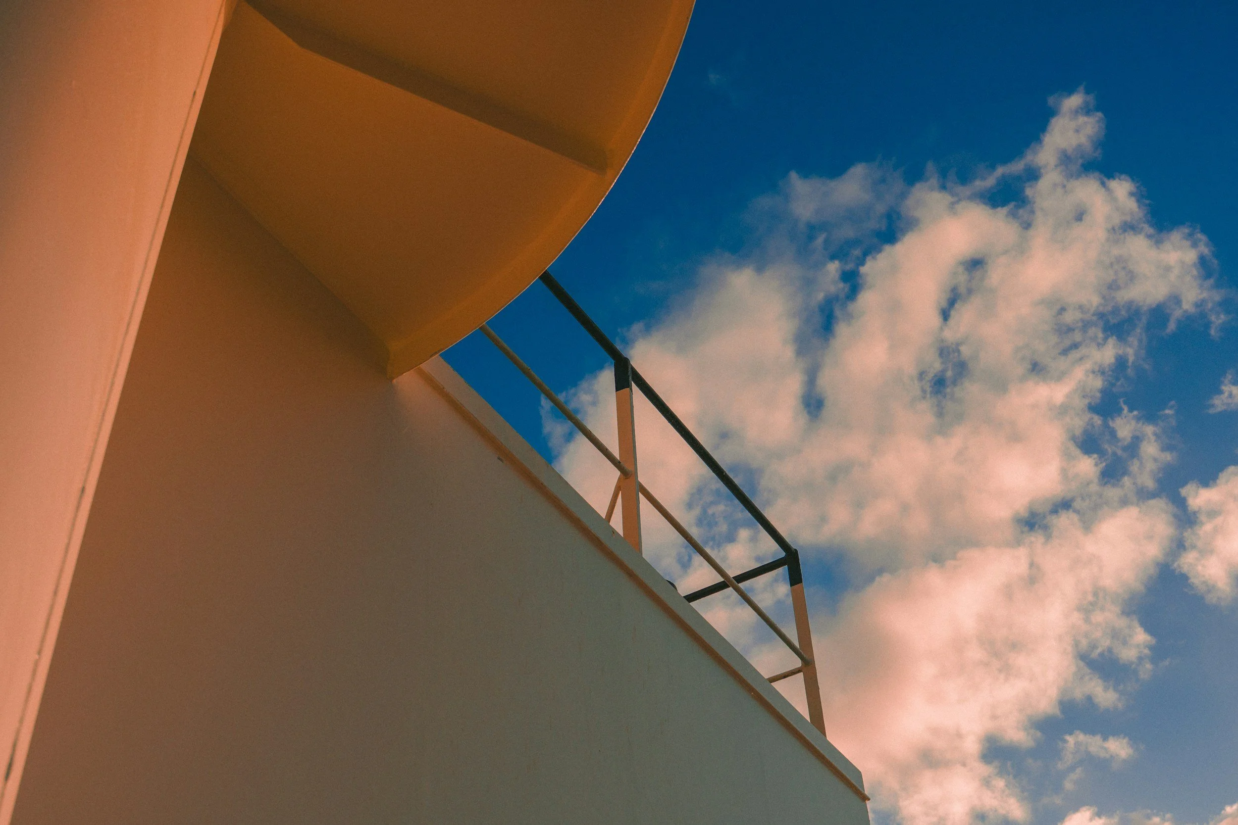 Looking up at a building's corner with a white wall, beige balcony, and black railing against a blue sky with white clouds.