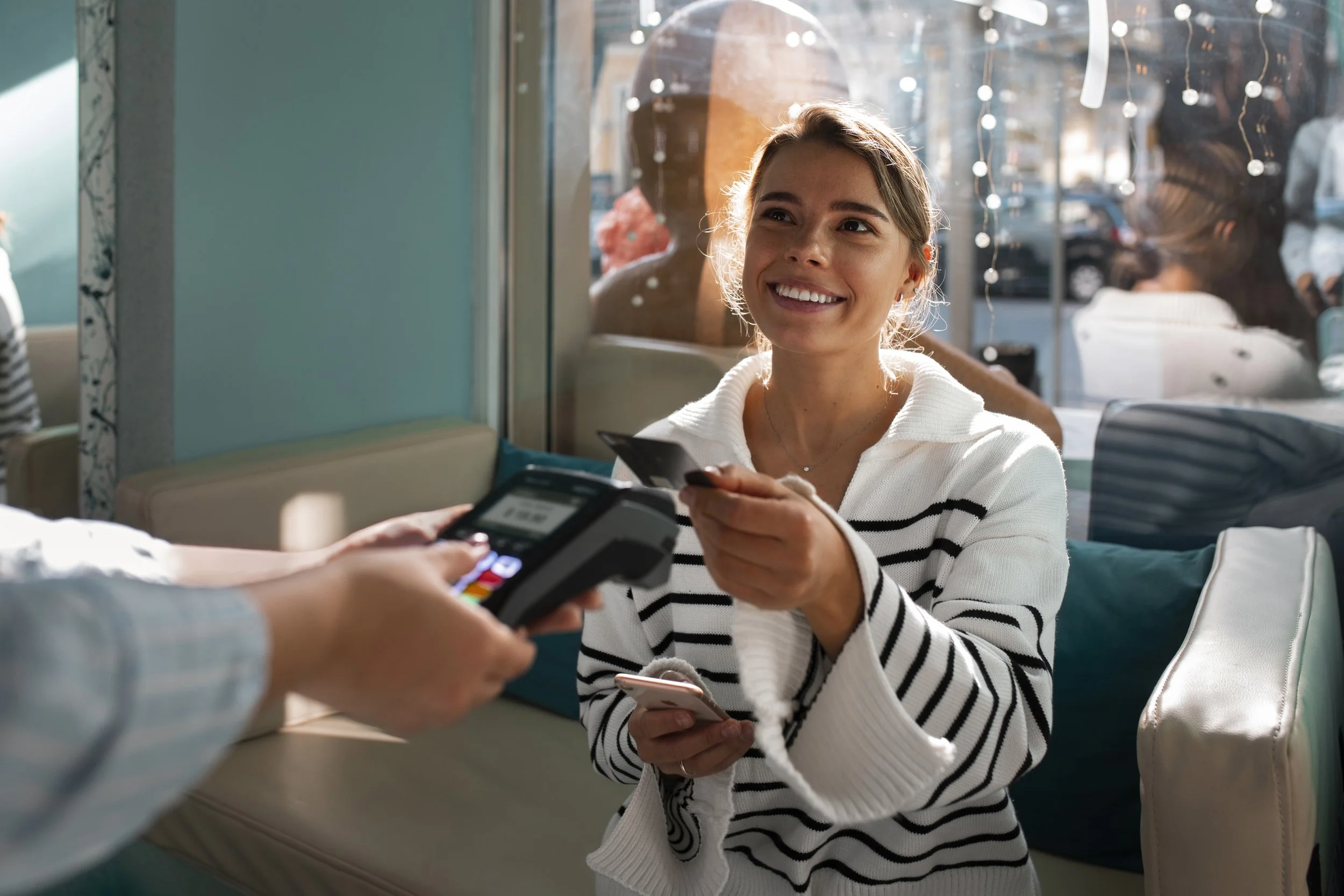 A woman in a white and black striped sweater is smiling while making a contactless payment with her card at a restaurant or cafe.
