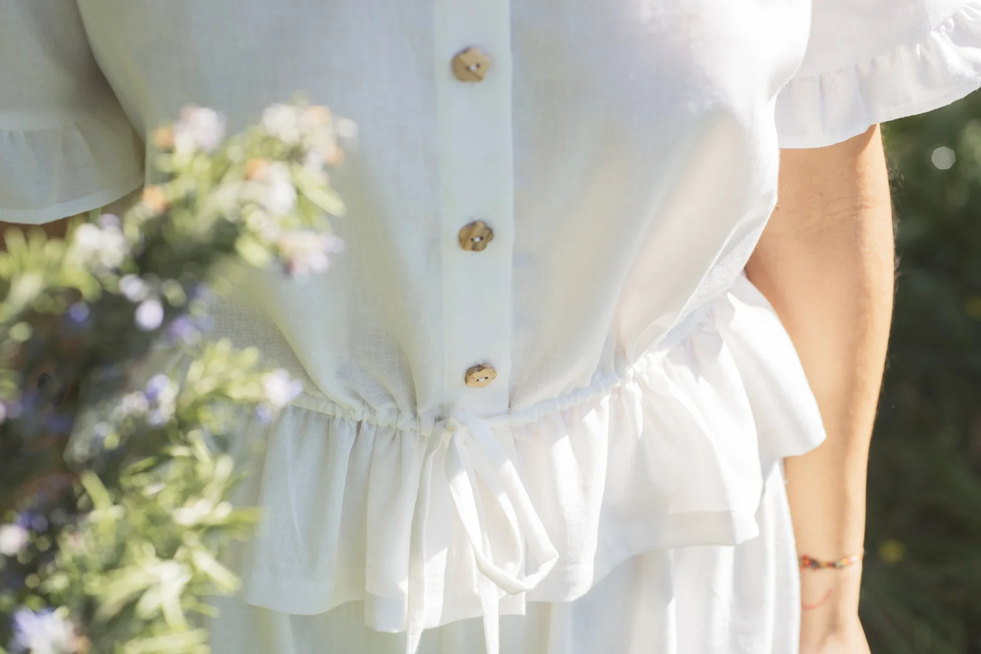 Blusa Aura de algodón orgánico en blanco, destacando sus botones de madera en forma de flor y el lazo ajustable en la cintura.