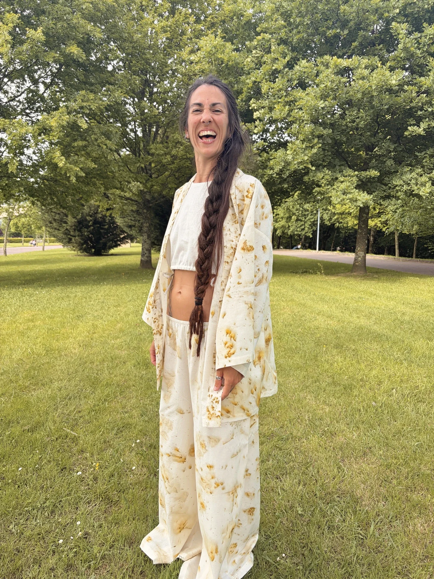 Mujer sonriendo en la naturaleza vistiendo un pantalón ancho y kimono a juego de algodón orgánico claro, teñido con tintes naturales,  con estampado botánico de flores doradas de Pericón.